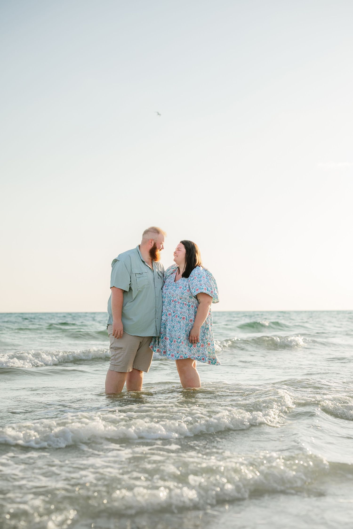 Couple embracing in ocean waves with soft sunset lighting and serene beach backdrop.