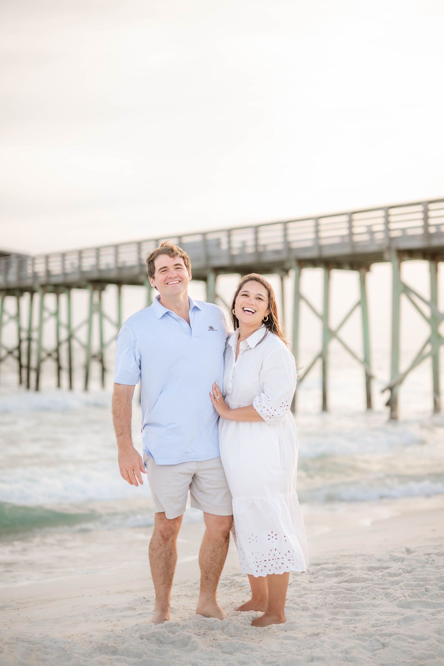 A joyful couple laughs together during their beach photoshoot near a pier.