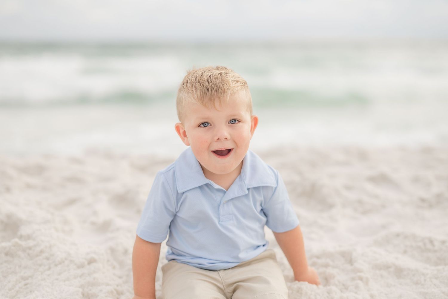 Child in blue polo shirt sitting on beach sand with ocean in background in Panama City Beach, Fl..