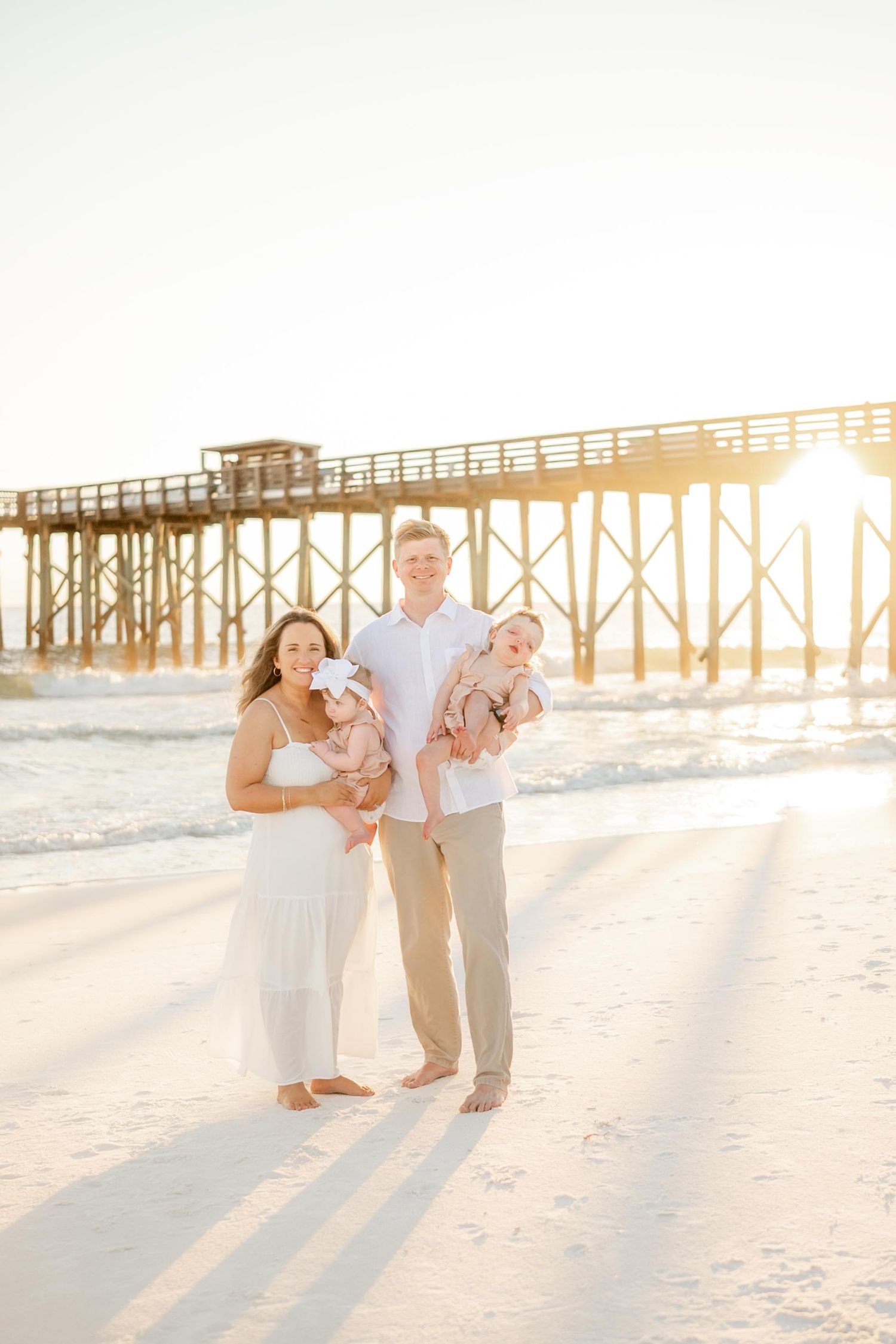 Family walking along beach with wooden pier and golden sunlight in background.