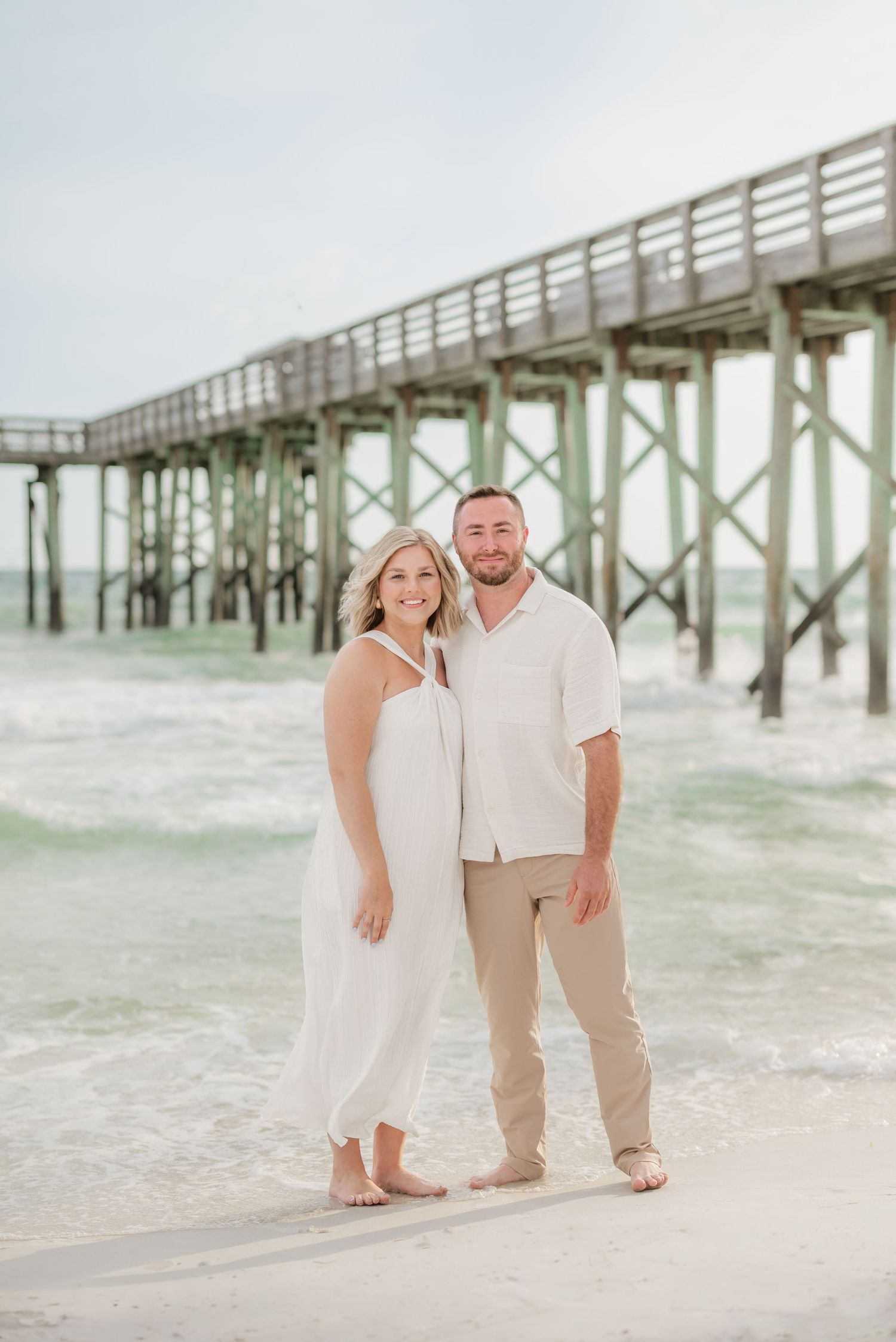 Couple in white and beige outfits posing near weathered beach pier.