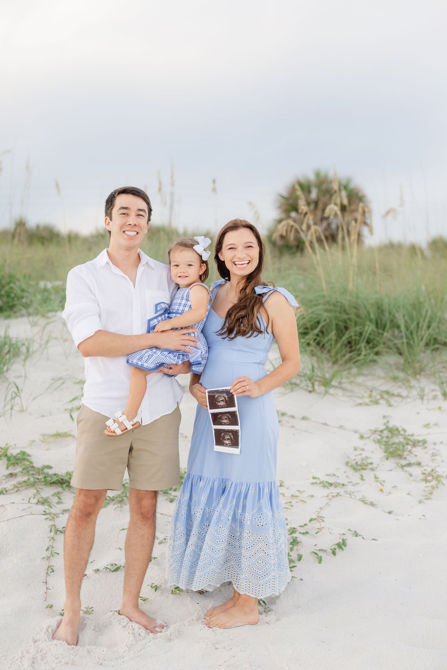 Family sharing a tender moment on sandy beach dunes with palm trees in background.