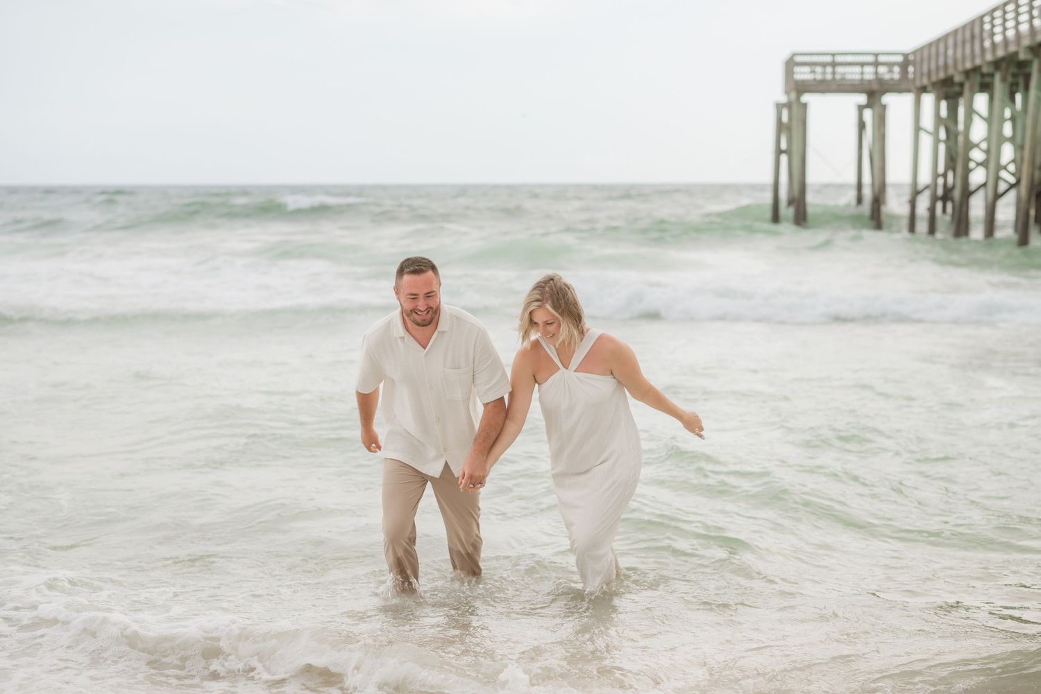 Couple splashing and playing in ocean waves near wooden pier.