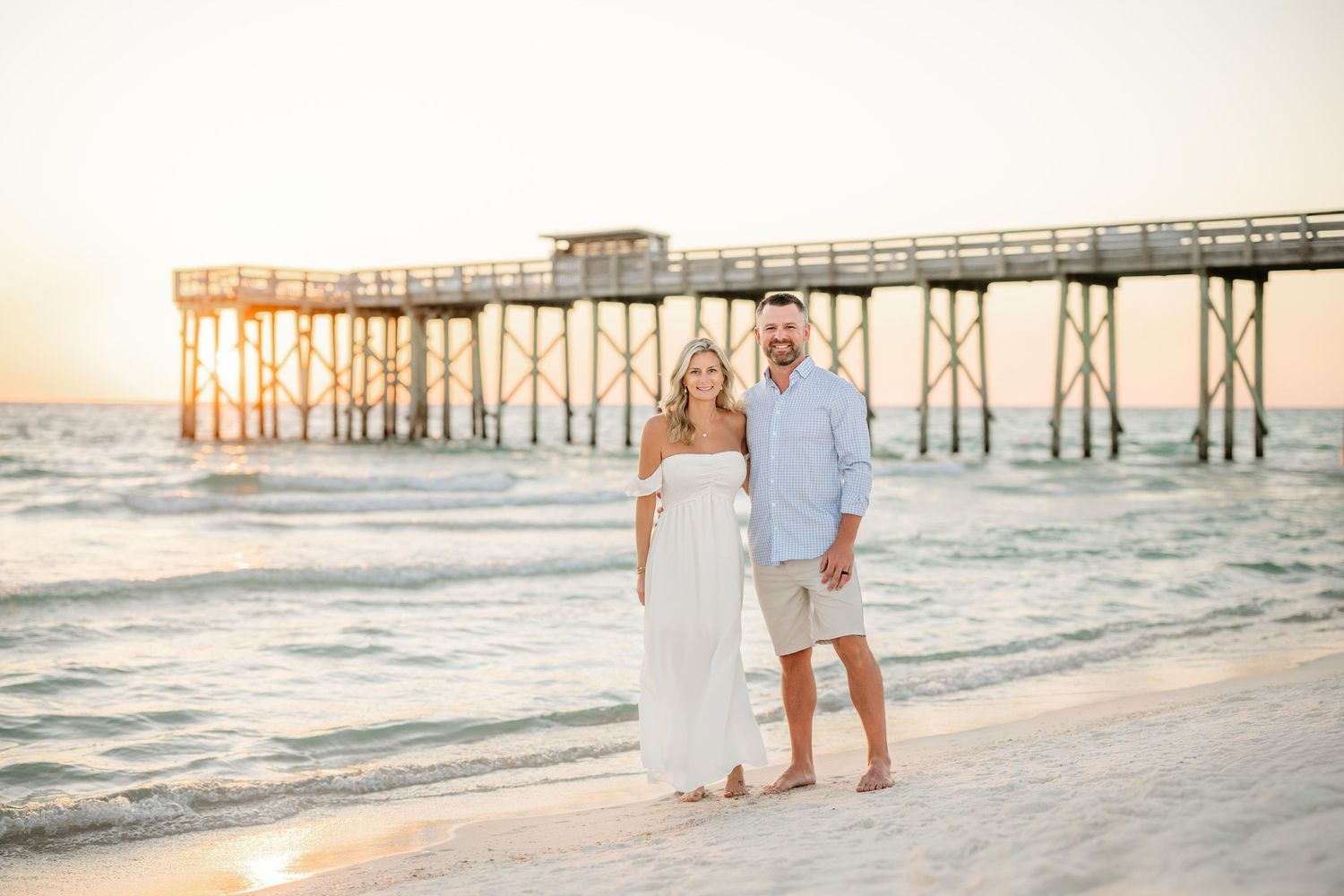 A couple poses on the beach at sunset with an old wooden pier in the background.
