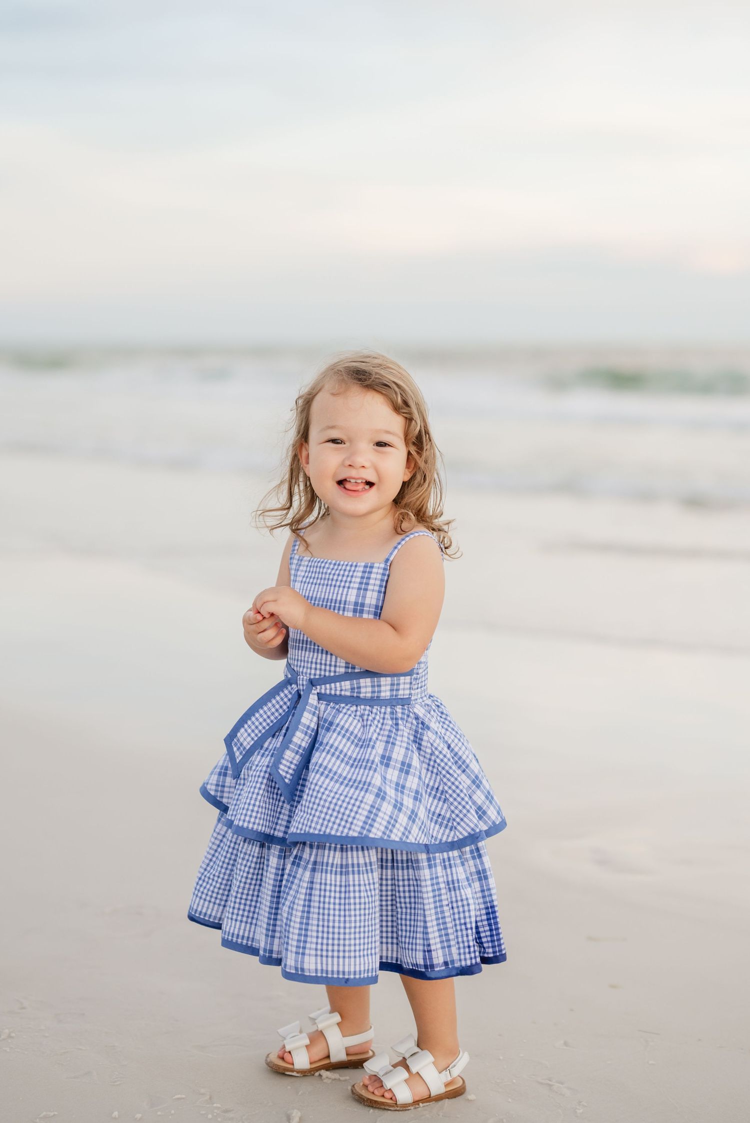 Toddler in blue ruffled dress and white sandals playing on beach.