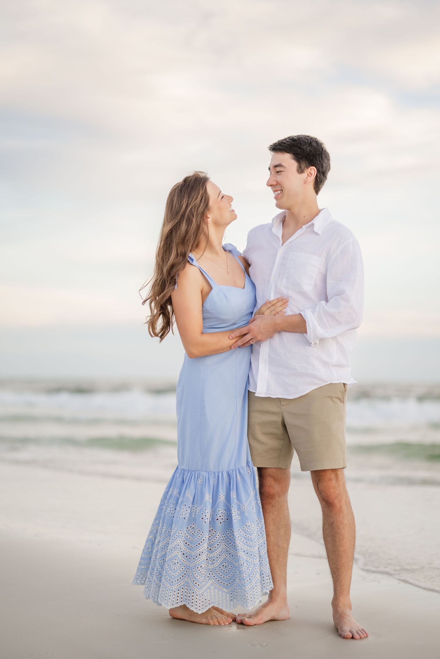 A romantic couple poses together on a sandy beach wearing blue and white outfits at dusk.