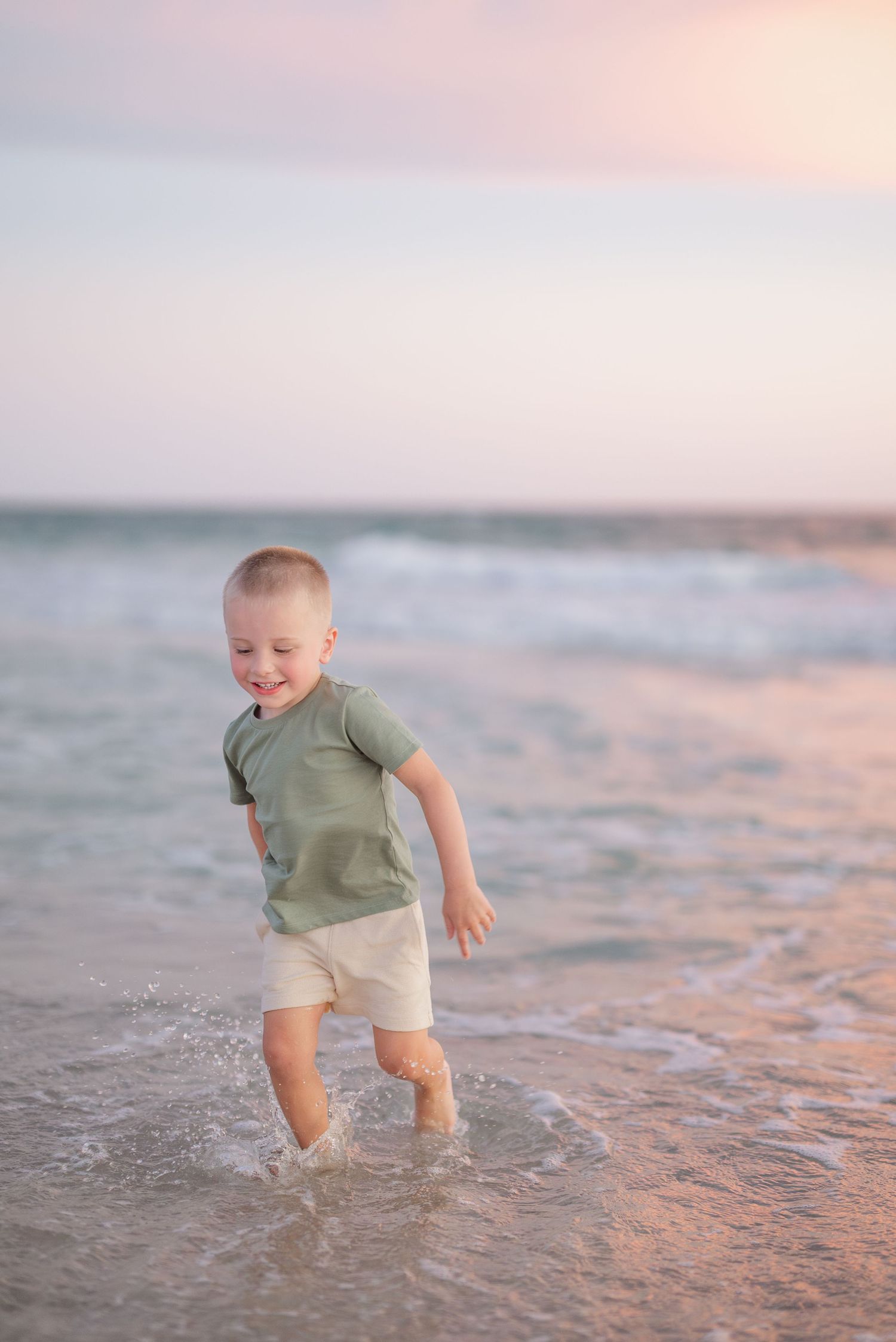 A child splashes playfully in shallow ocean waves while wearing a green t-shirt and khaki shorts.