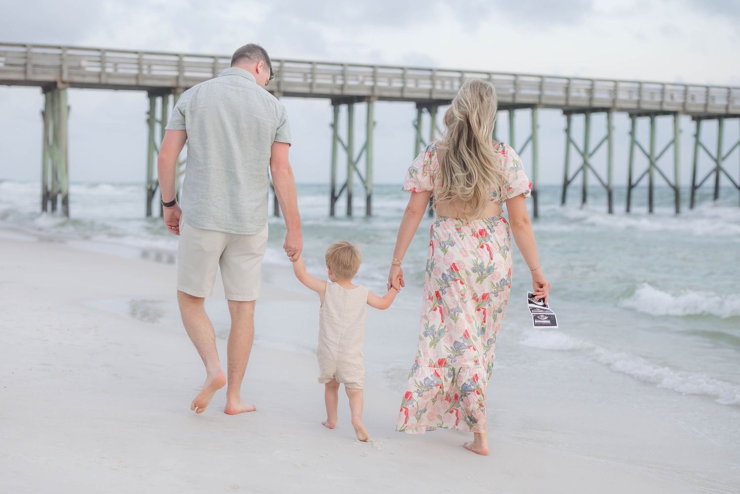 A family walks along the shoreline with a wooden pier visible in the distance.