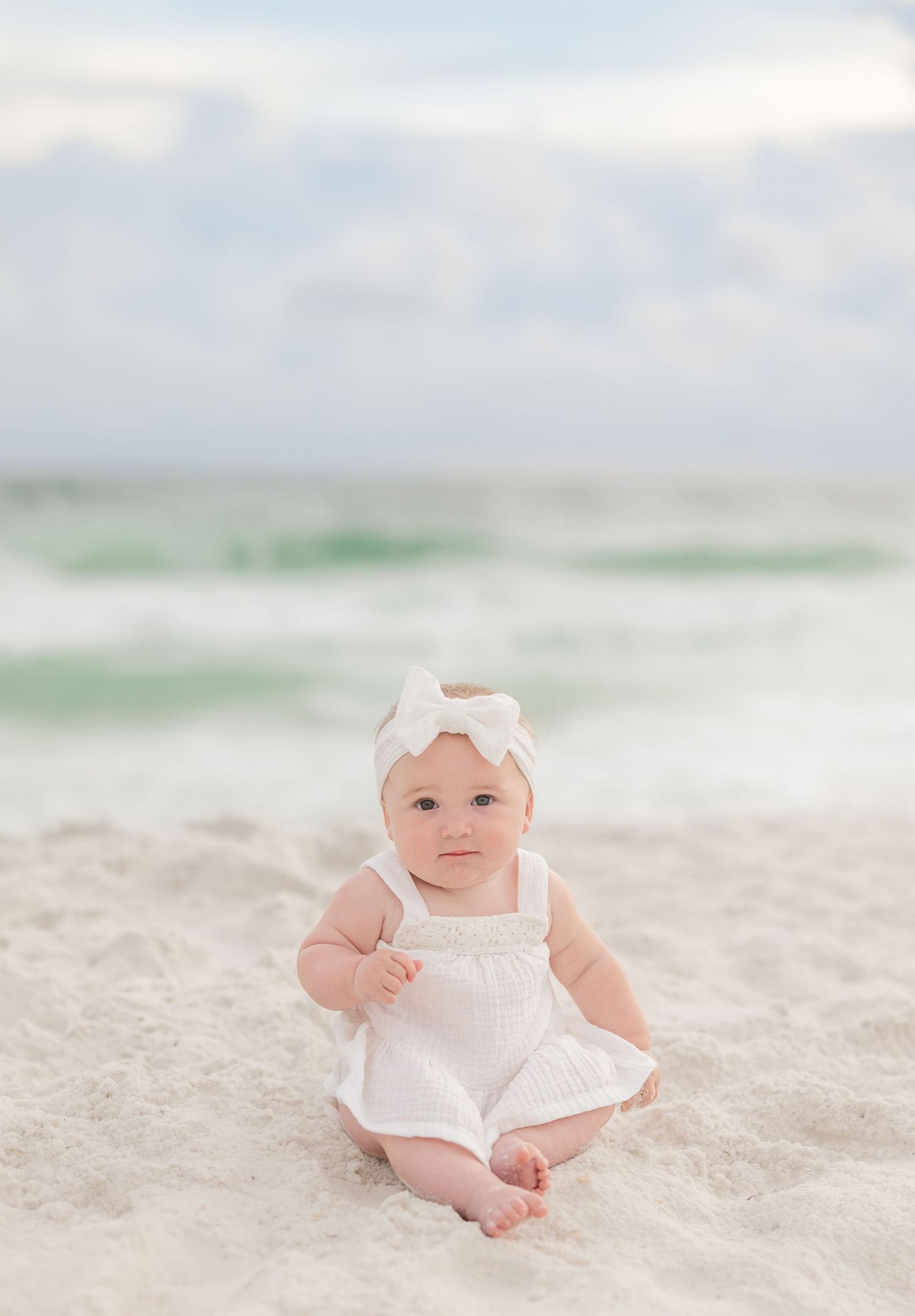 A baby wearing a white dress and bow headband sits on a sandy beach with soft pastel colors in the background in Panama City Beach, Fl..