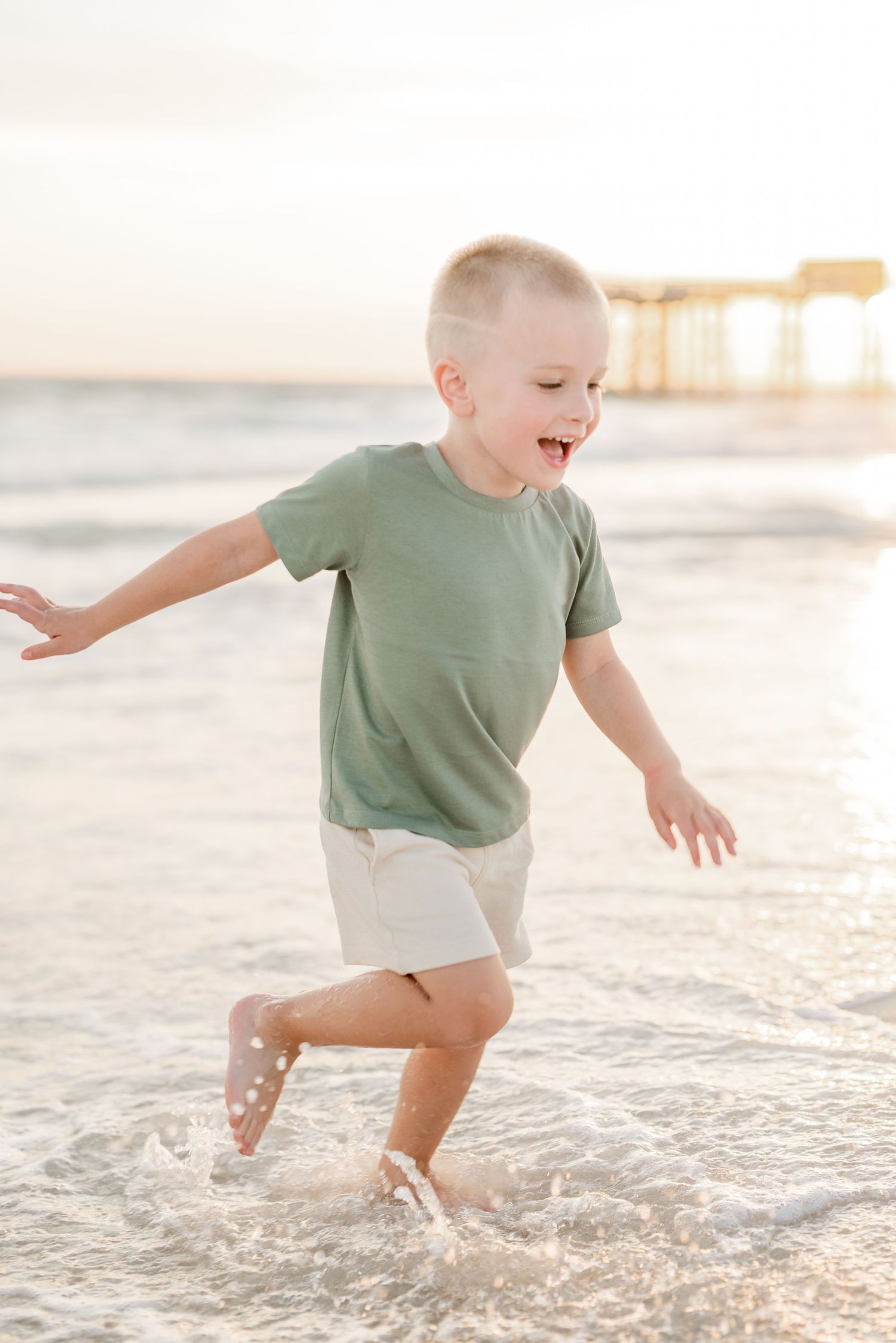 Child in green shirt and white shorts splashing in ocean waves at sunset.