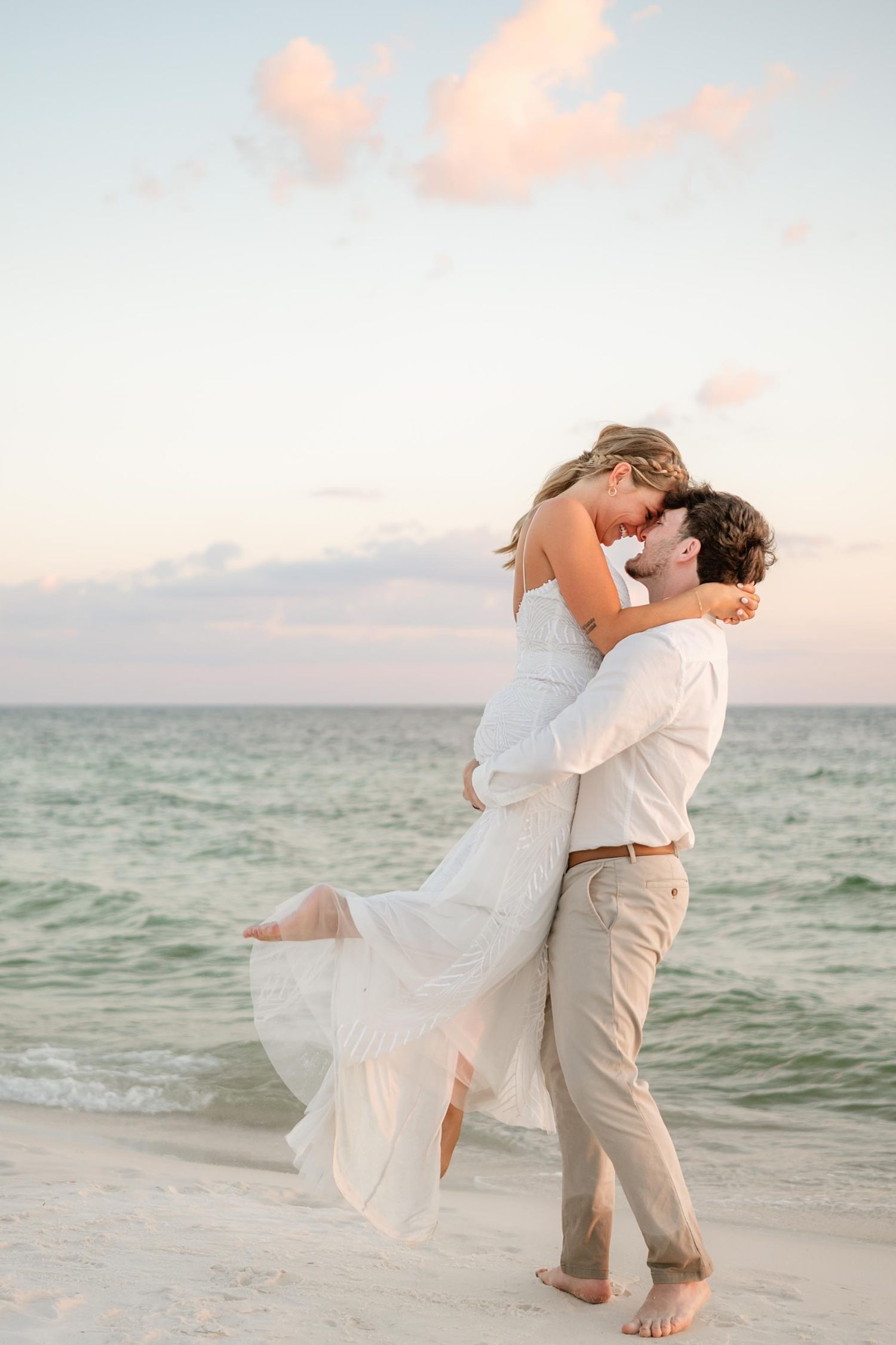 A joyful couple embraces on a beach at sunset with the bride's dress flowing in the breeze.