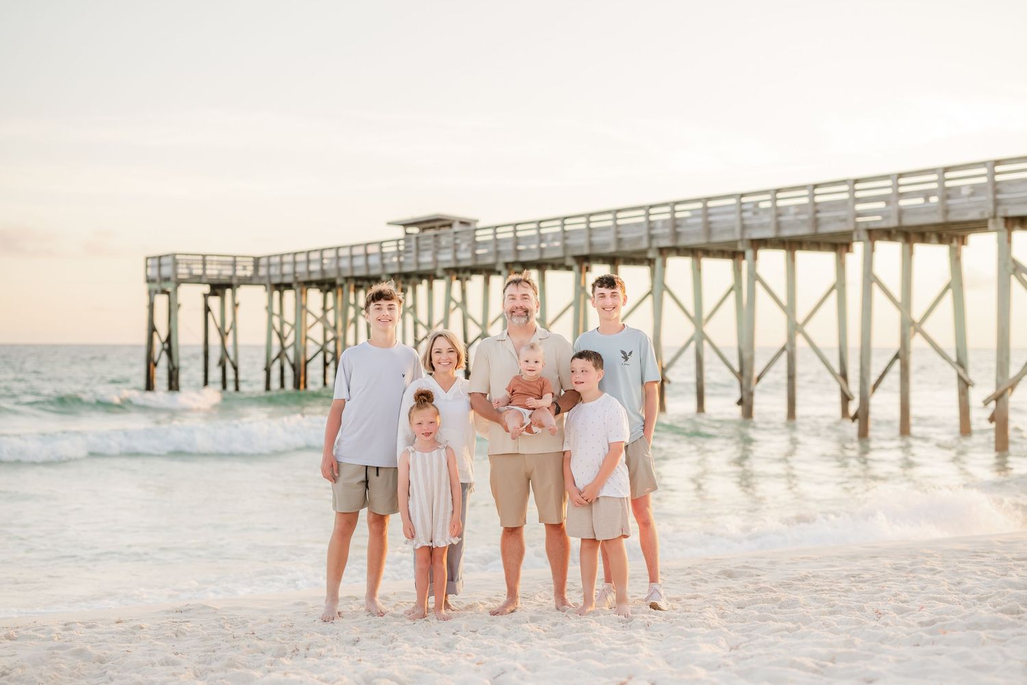 Family walking together on white sandy beach near wooden pier during sunset photoshoot.