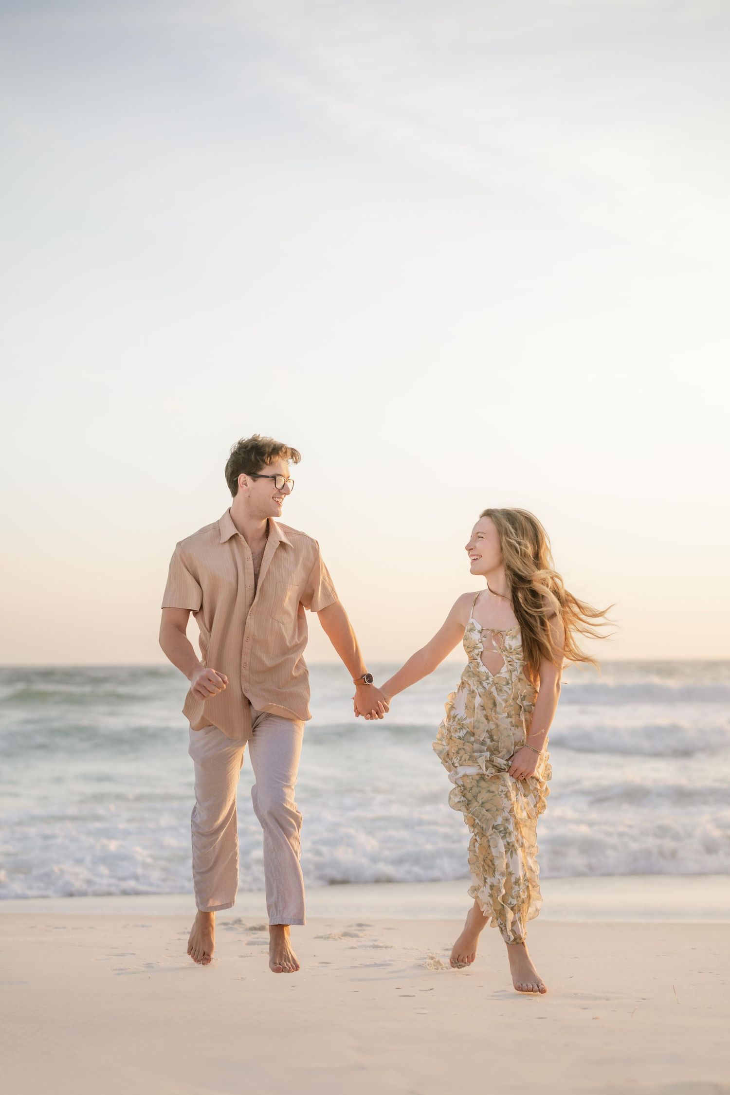 A couple holds hands and walks barefoot on a sandy beach at sunset, wearing coordinating neutral and floral outfits.