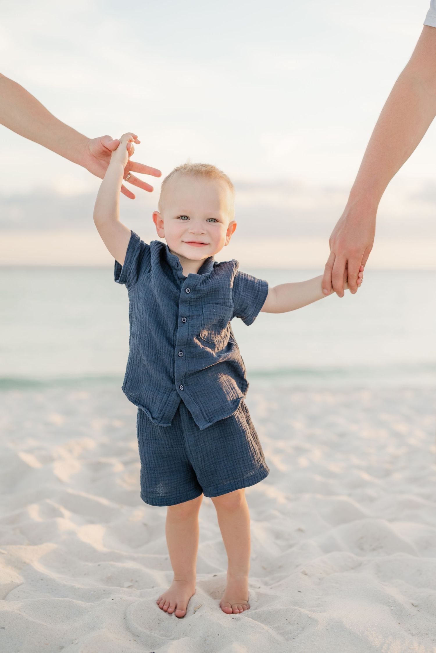 A toddler takes first steps on white sand beach while being supported by adult hands.