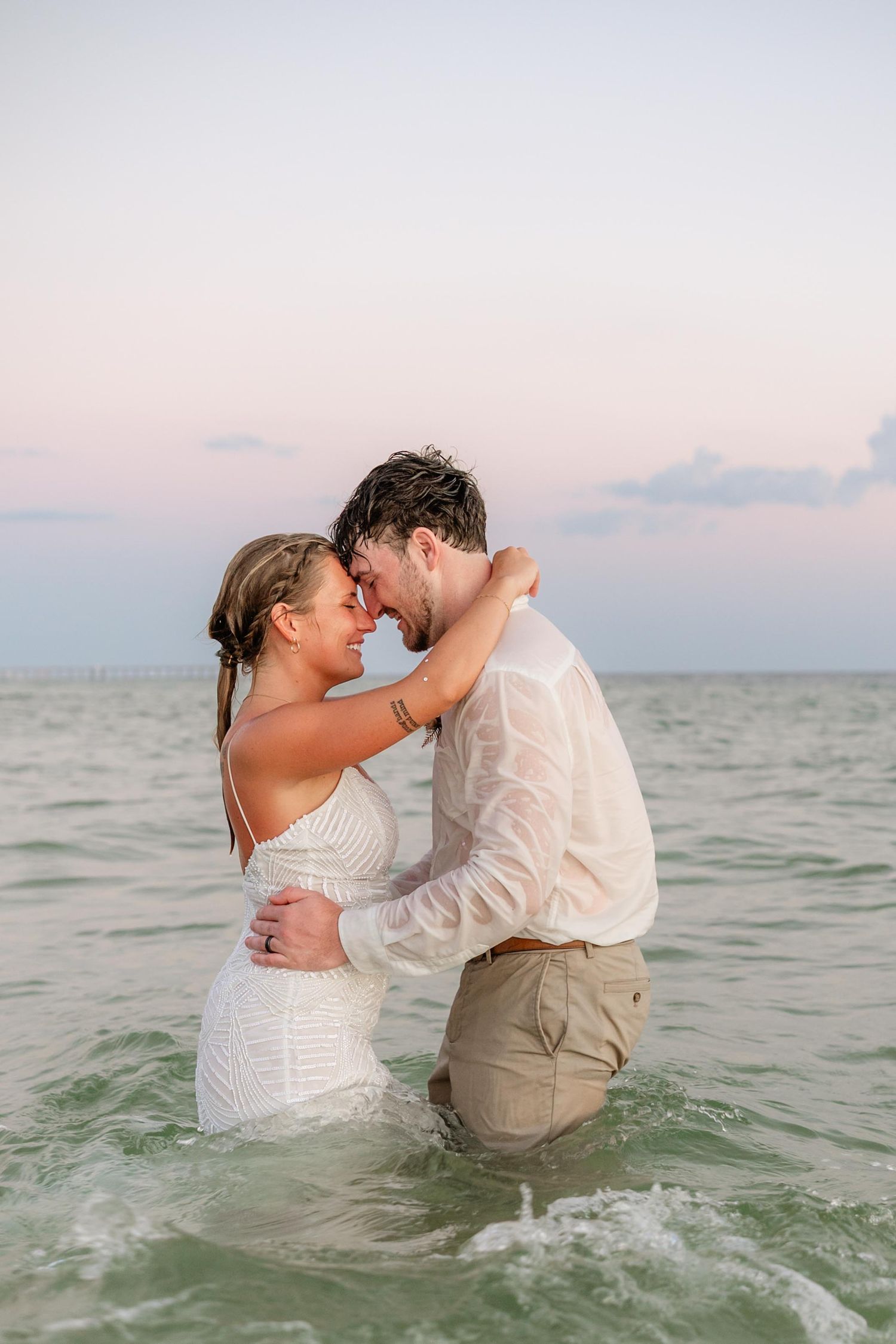 Romantic couple embraces while standing in ocean water during sunset photoshoot.