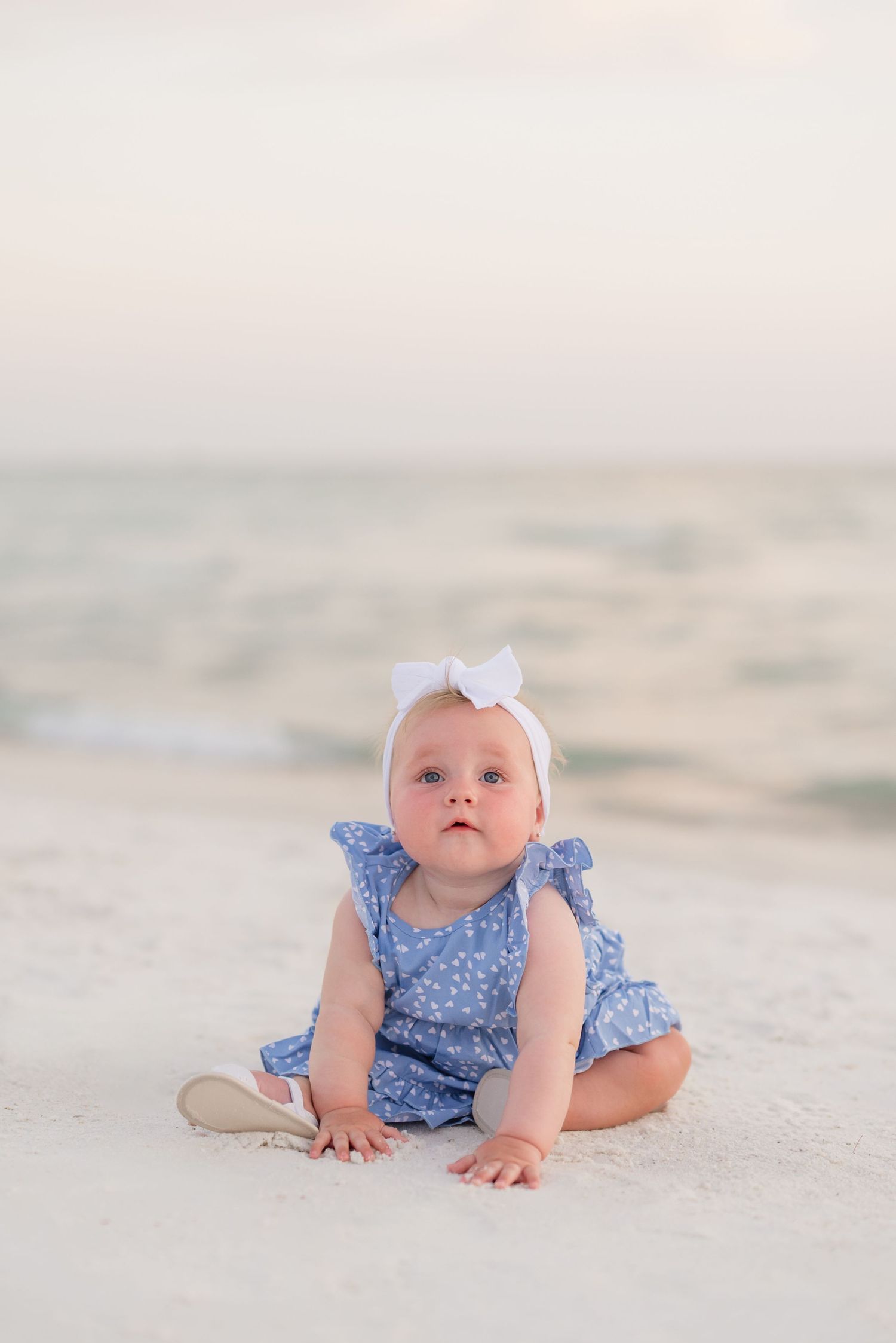 A baby in blue floral outfit and white bow sits on sandy beach during sunset.