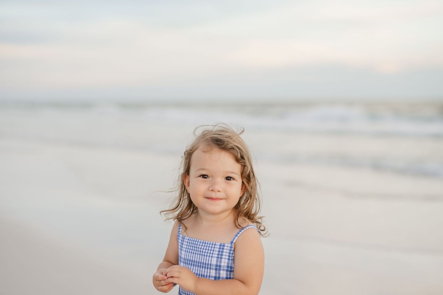 Child in blue and white gingham dress standing on white sandy beach.