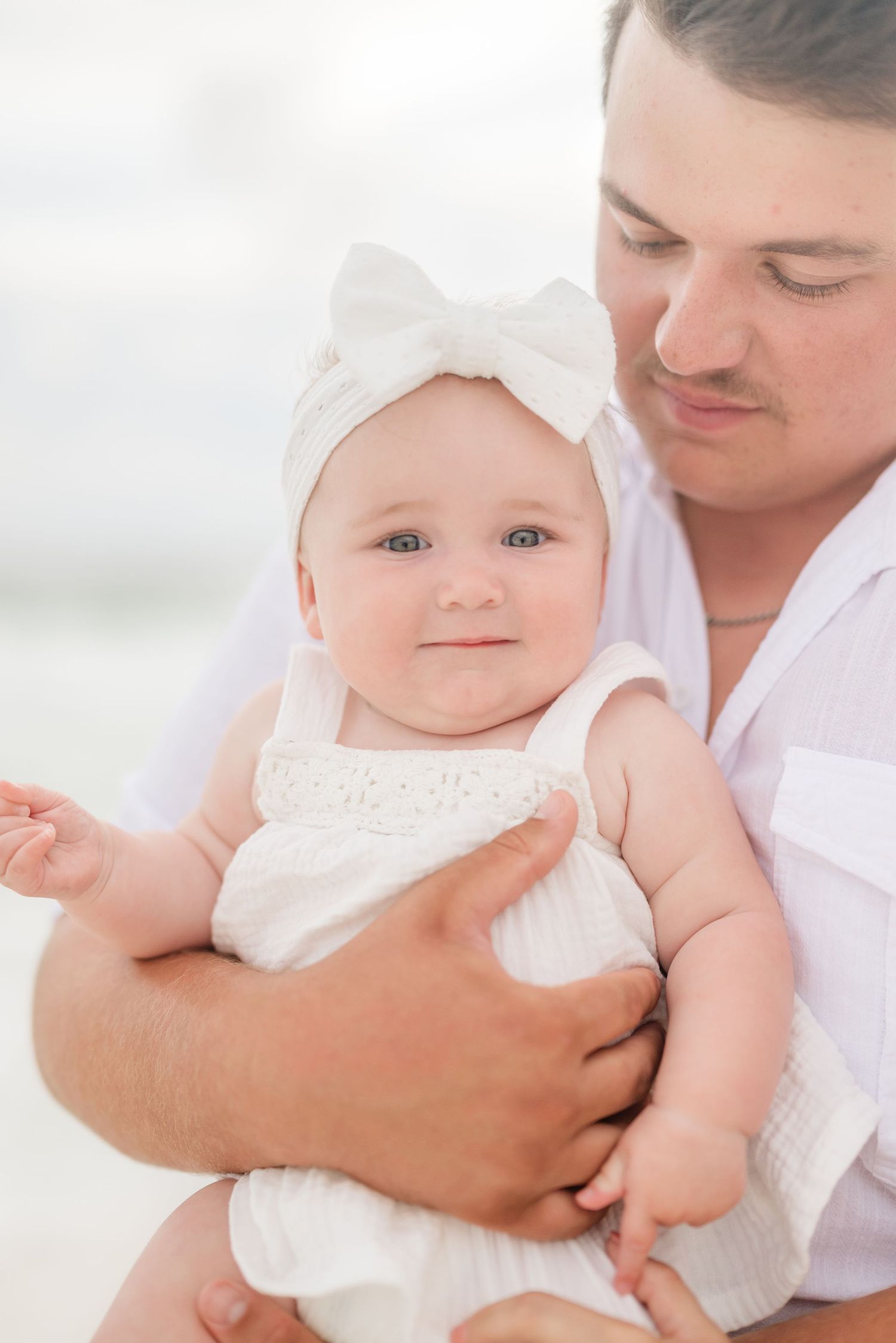 Baby wearing white outfit held lovingly at beach photoshoot.