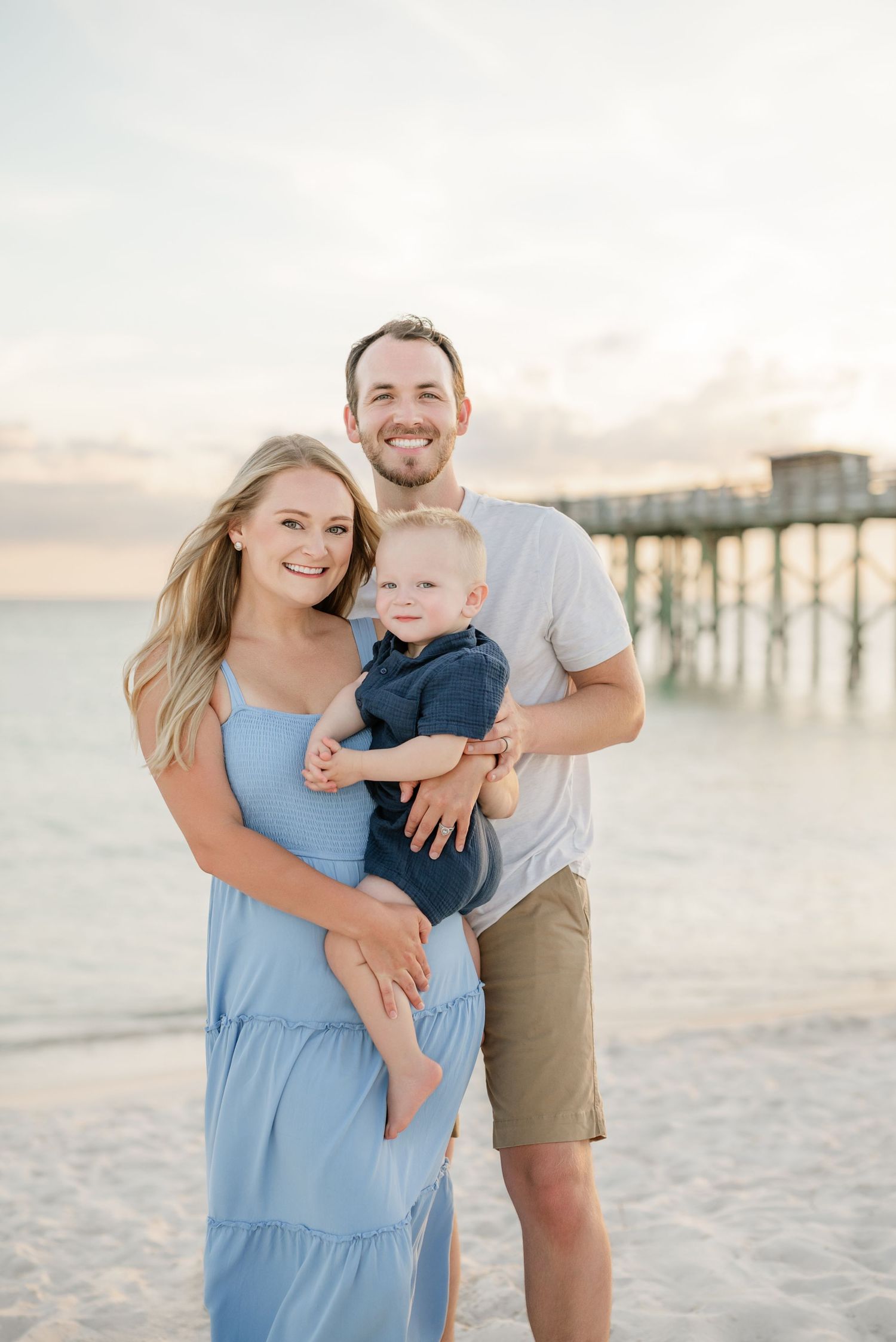 Family enjoying sunset at beach pier with soft lighting and sandy backdrop in coordinated blue and tan outfits.