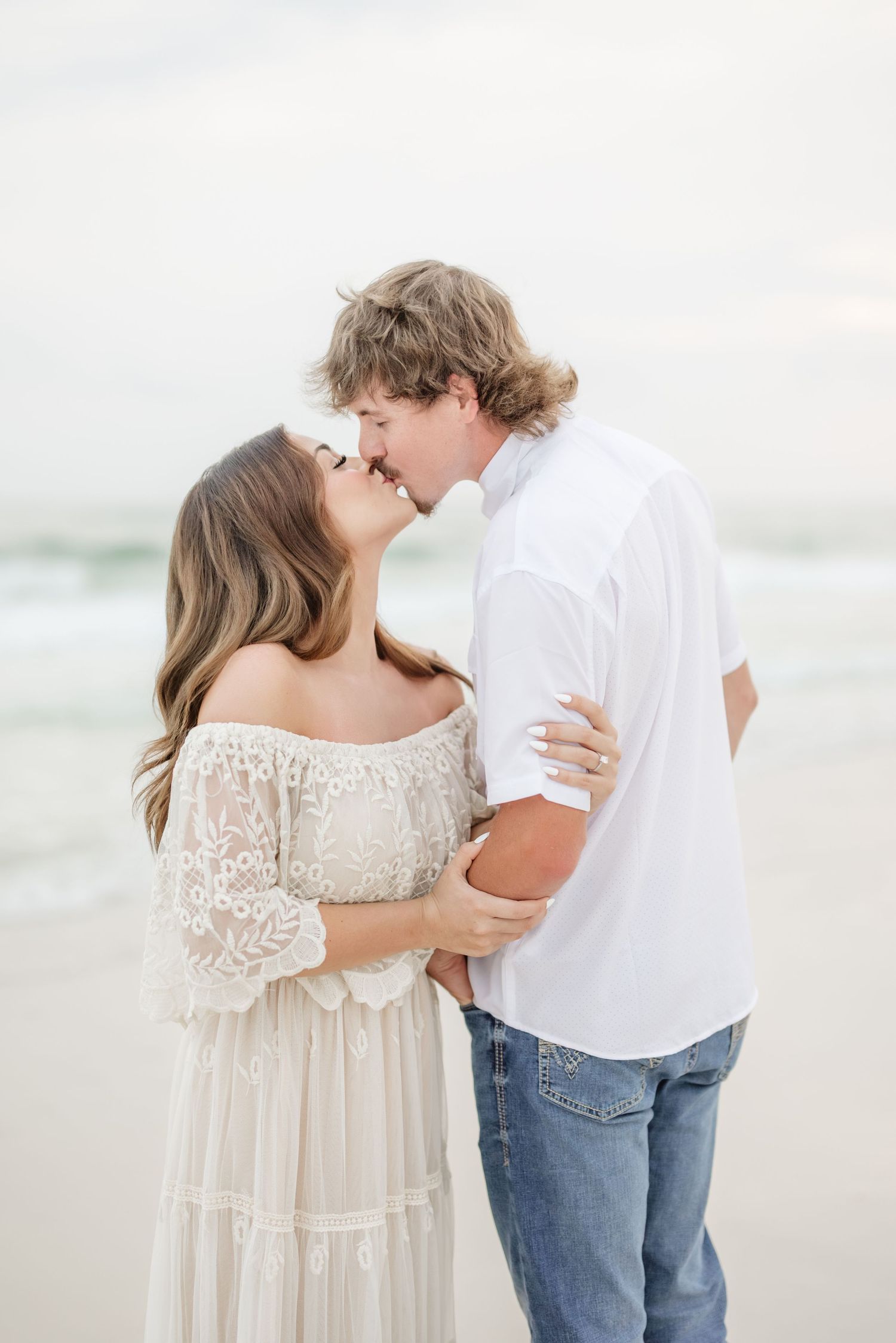 Romantic beach engagement photos with couple in white and denim sharing tender moments.