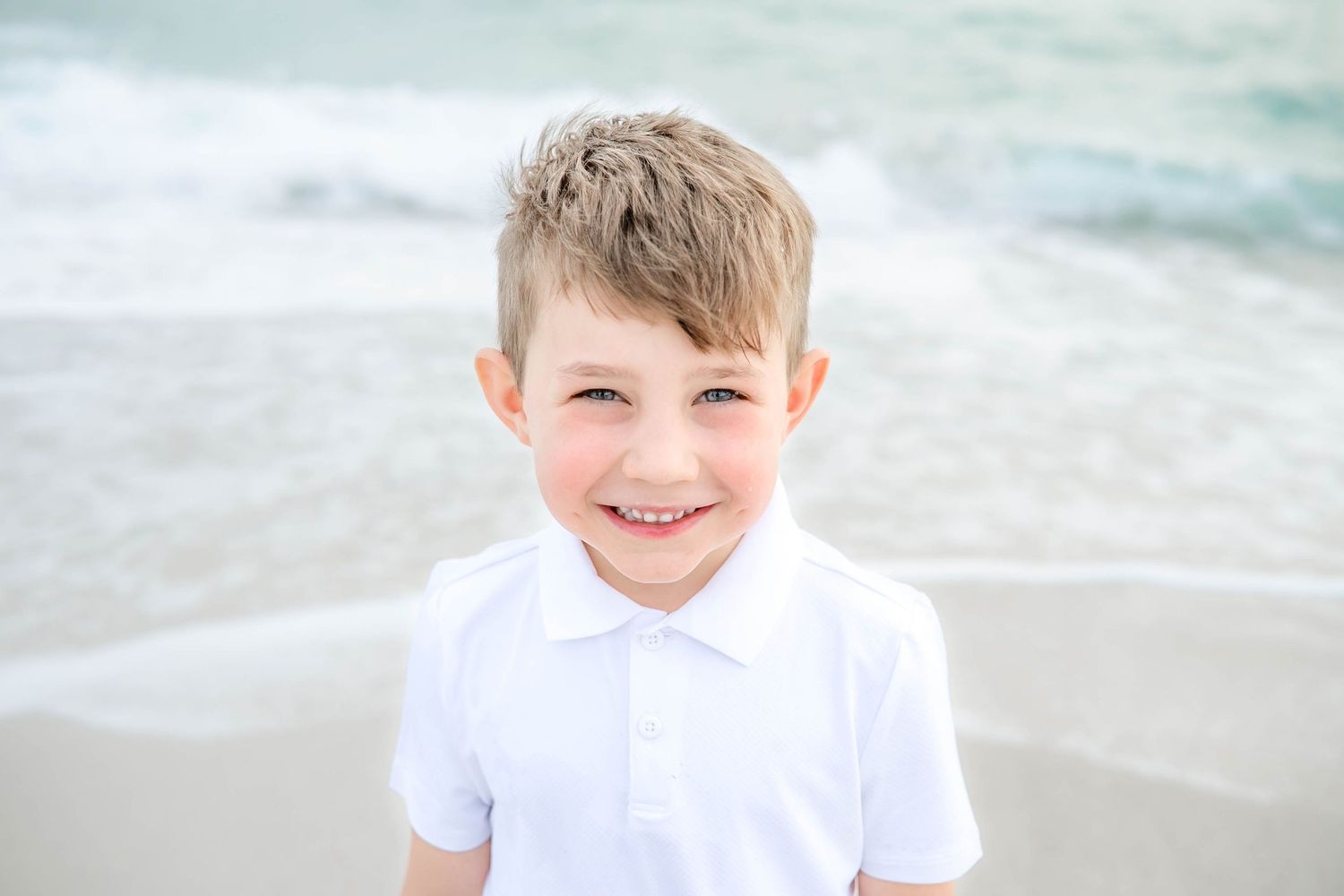 Young person in white shirt smiling on a sunny beach with ocean waves in background.