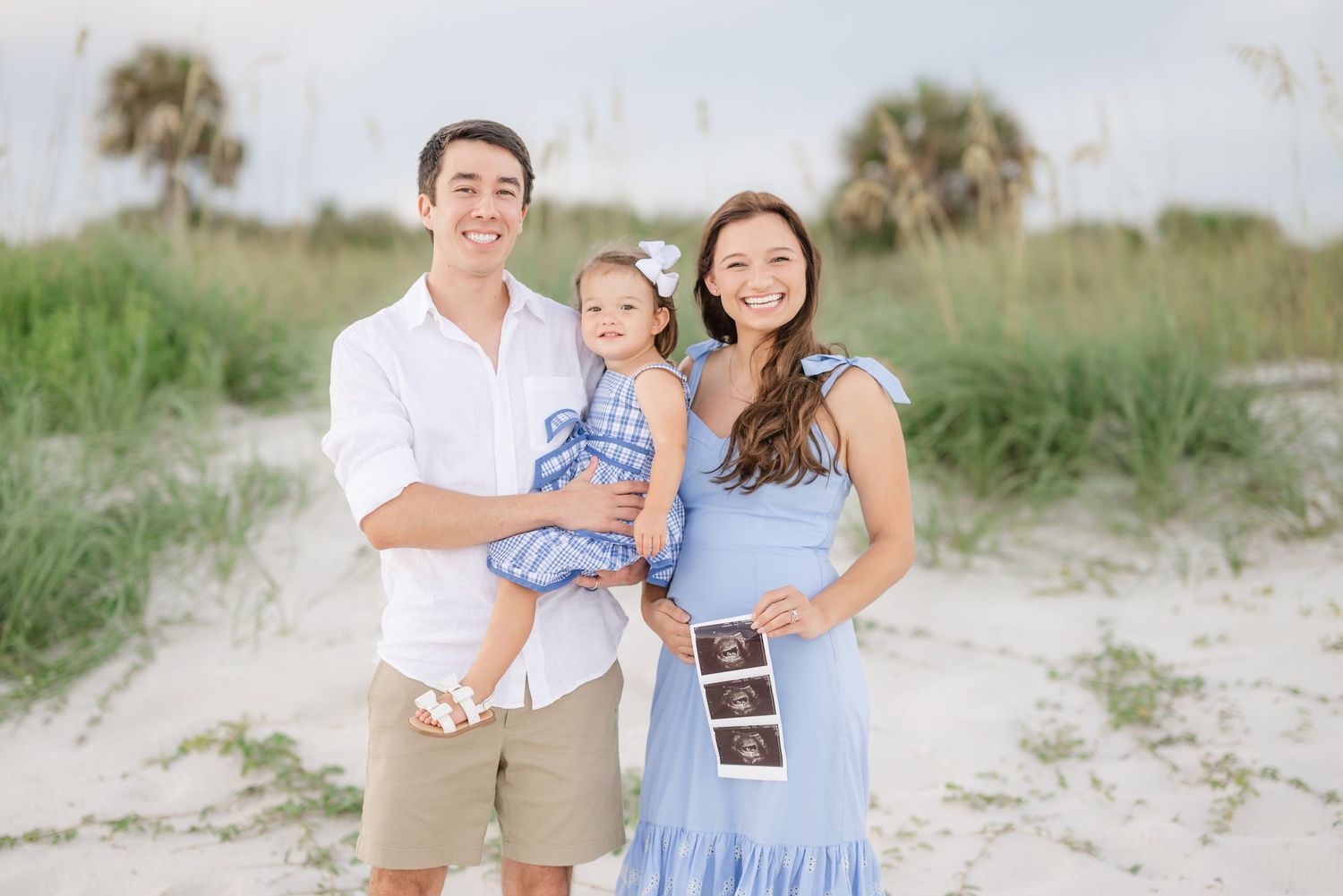 Family in coordinating blue and white outfits posing together on beach dunes at sunset.