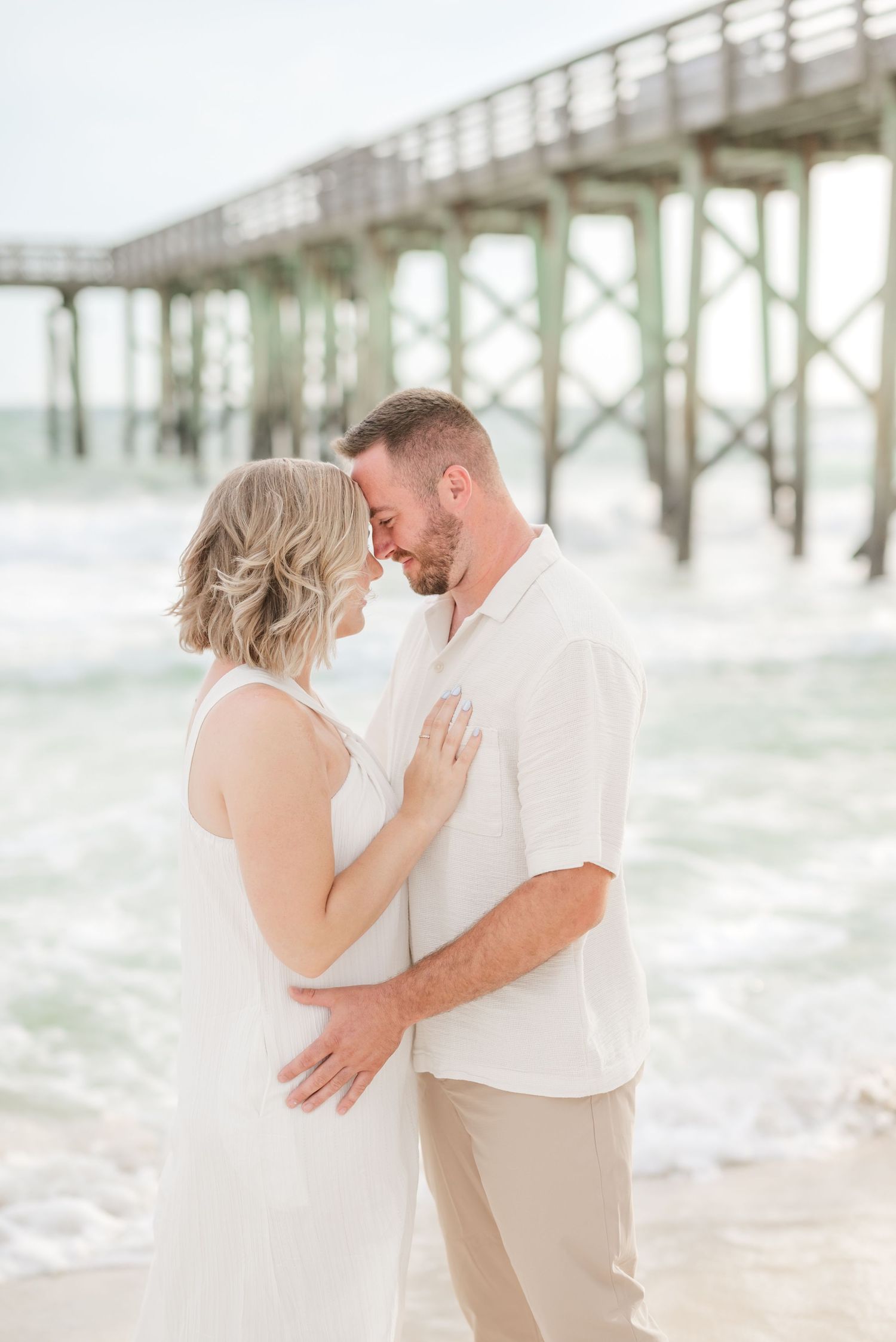 A romantic couple embraces on a beach with a wooden pier in the background.
