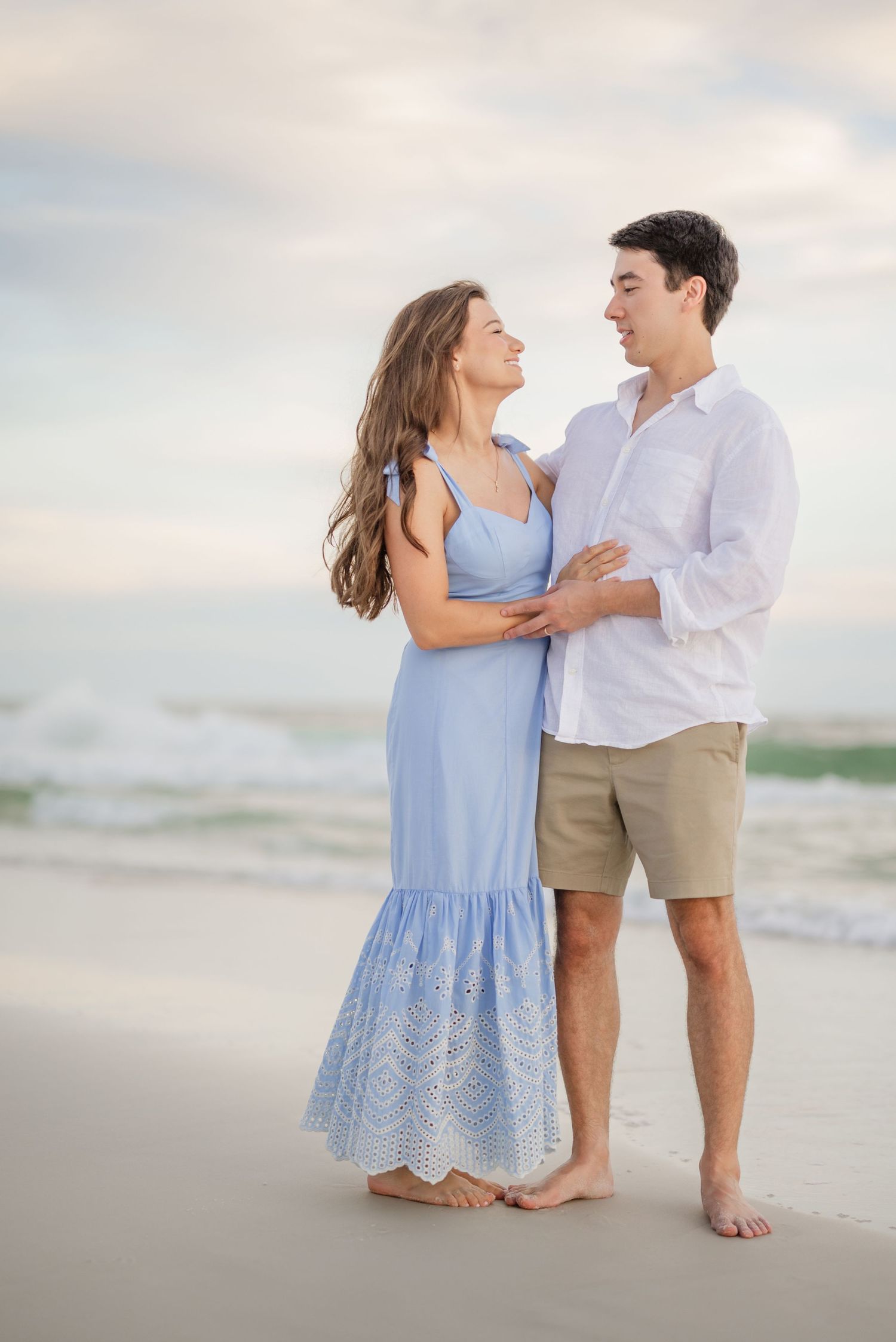 A couple stands barefoot in the sand during a beach photoshoot at dusk.