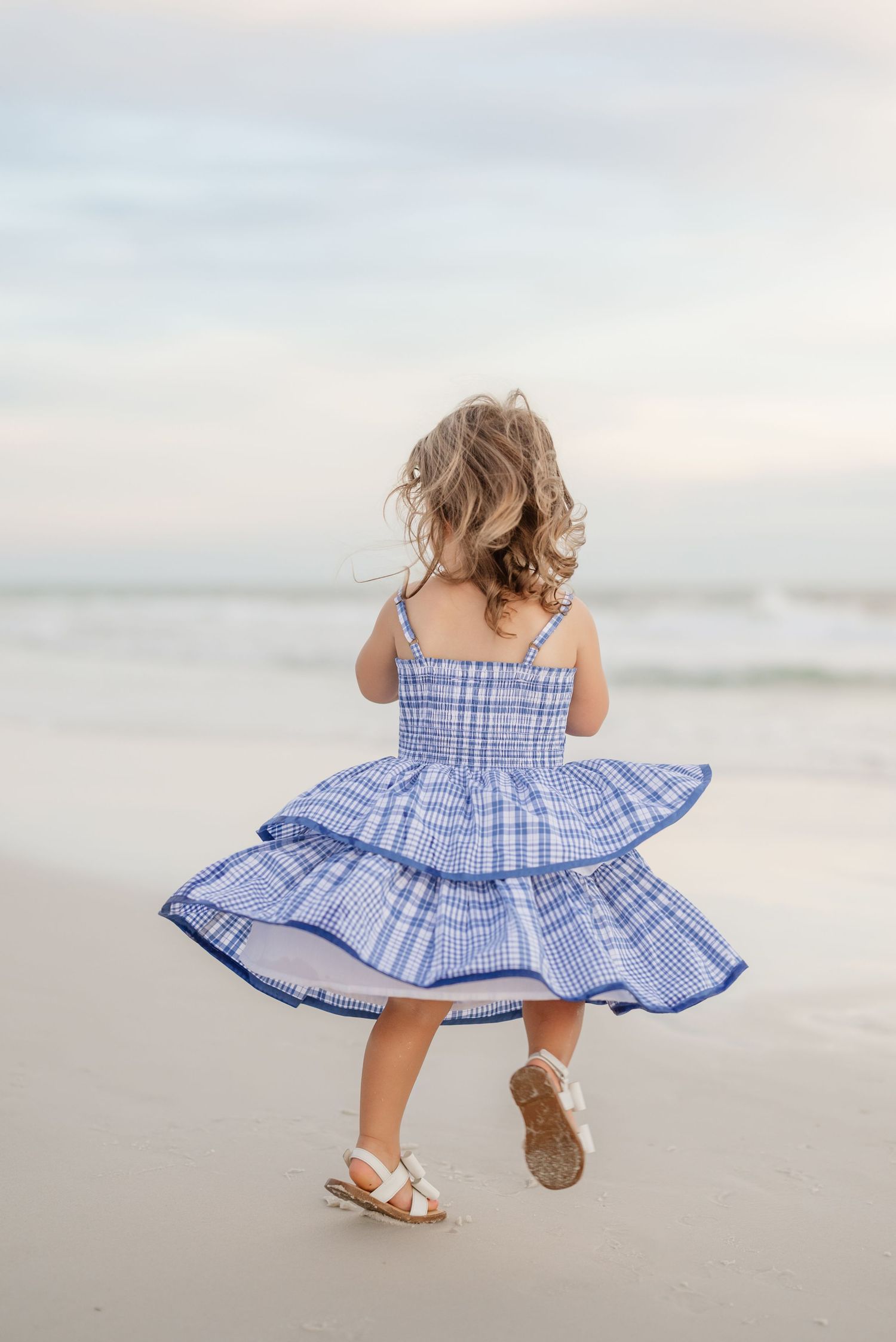Back view of child in blue ruffled dress twirling on beach at sunset.