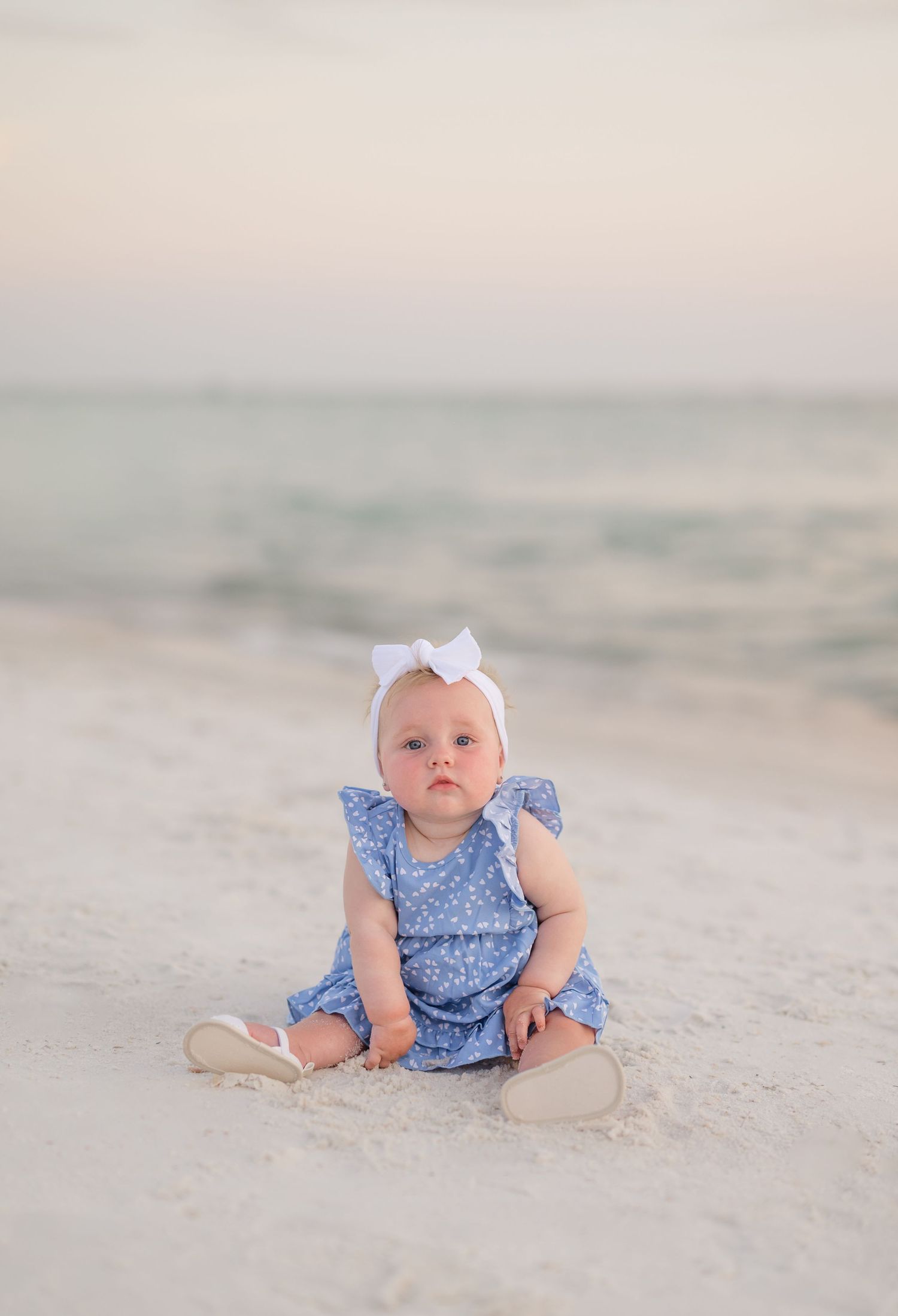 A baby in a blue floral dress and white bow sits on a peaceful beach at sunset.