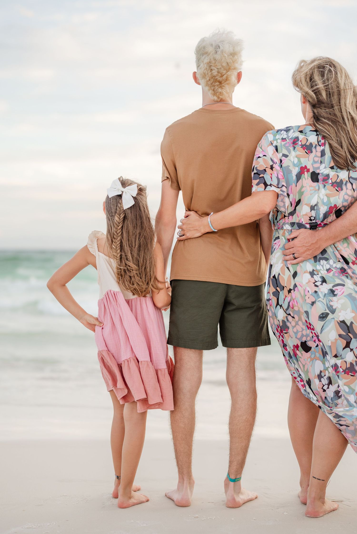 Back view of multi-generational family members standing together on beach facing ocean waves.