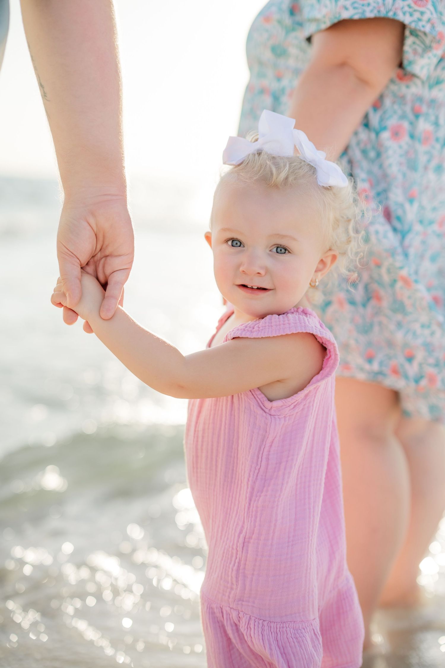 A toddler in a pink dress plays at the beach's edge with sparkling water in the background.
