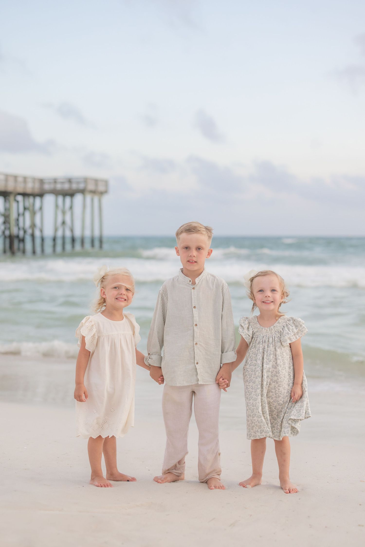 Children in light colored outfits standing together on beach with pier in background.