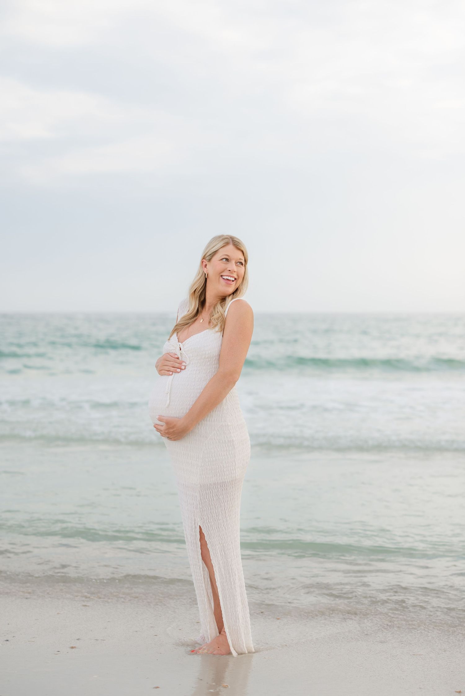 Maternity photo of woman in white dress standing on beach at sunset with ocean waves in background in Panama City Beach, Fl..
