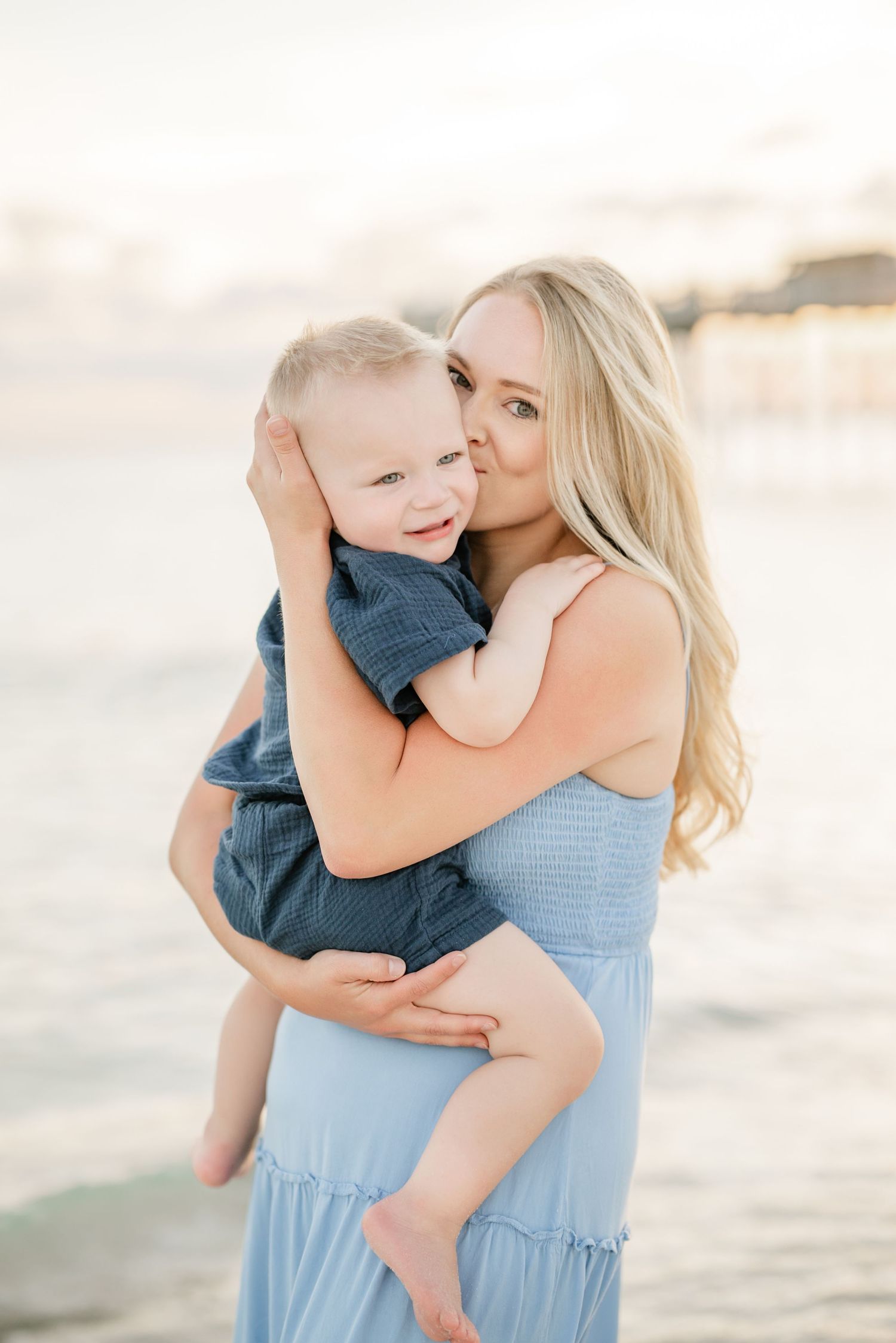 A blonde mother in a light blue dress holds and kisses her smiling toddler boy on a sunny beach.
