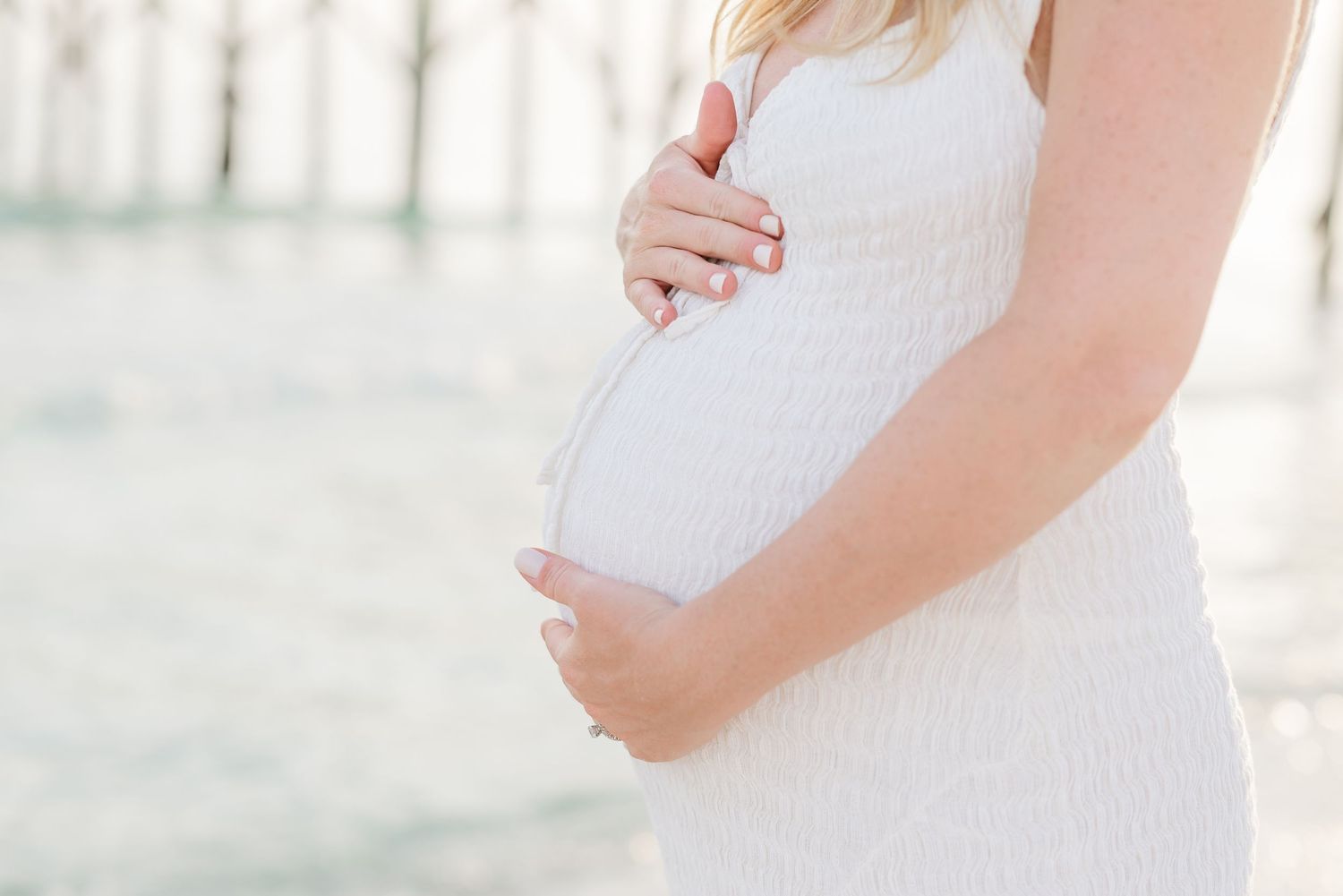 Close-up shot of pregnant belly in white lace dress against beach backdrop in Panama City Beach, Fl..