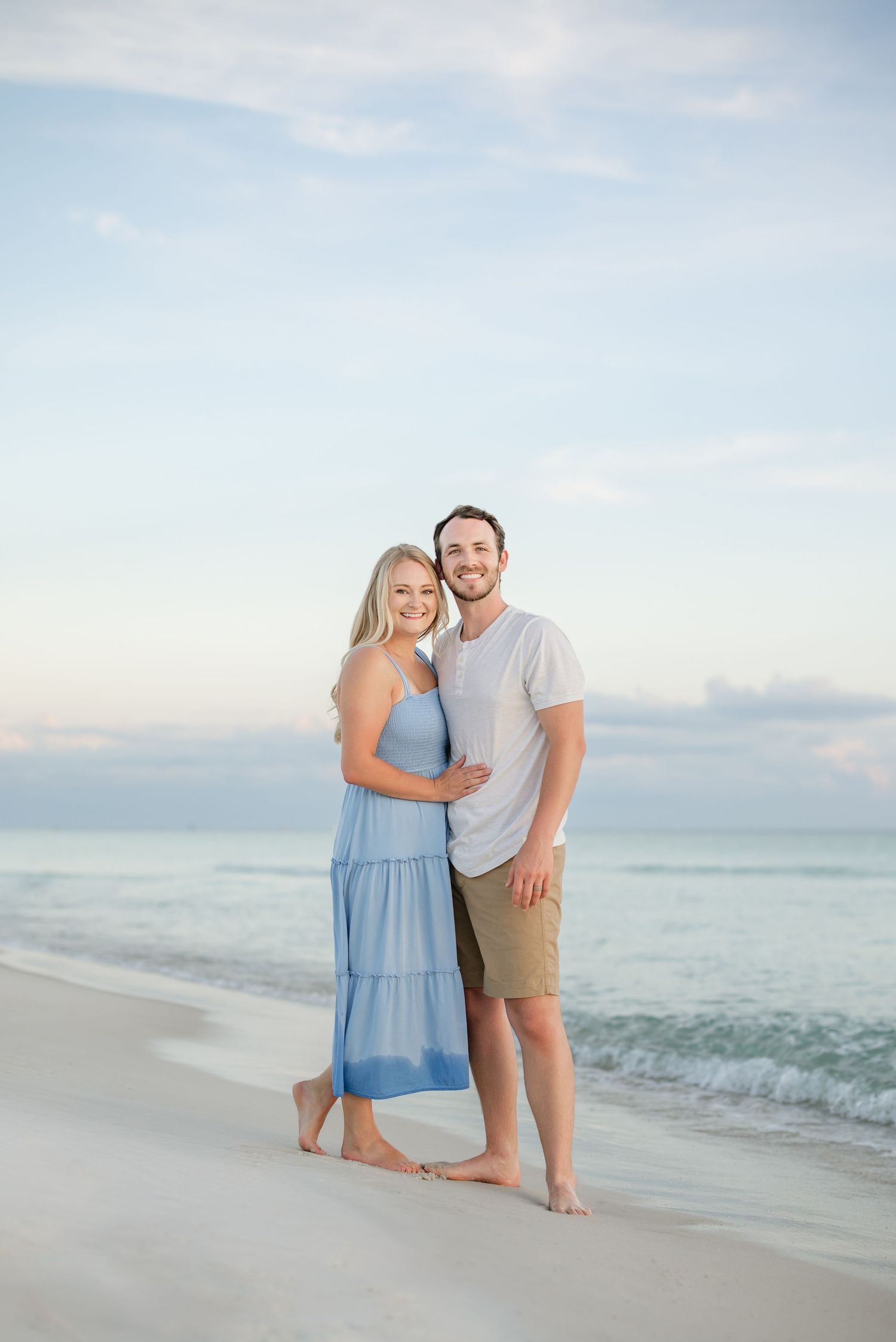 Loving couple poses on sandy beach with calm ocean and pastel sky behind them.