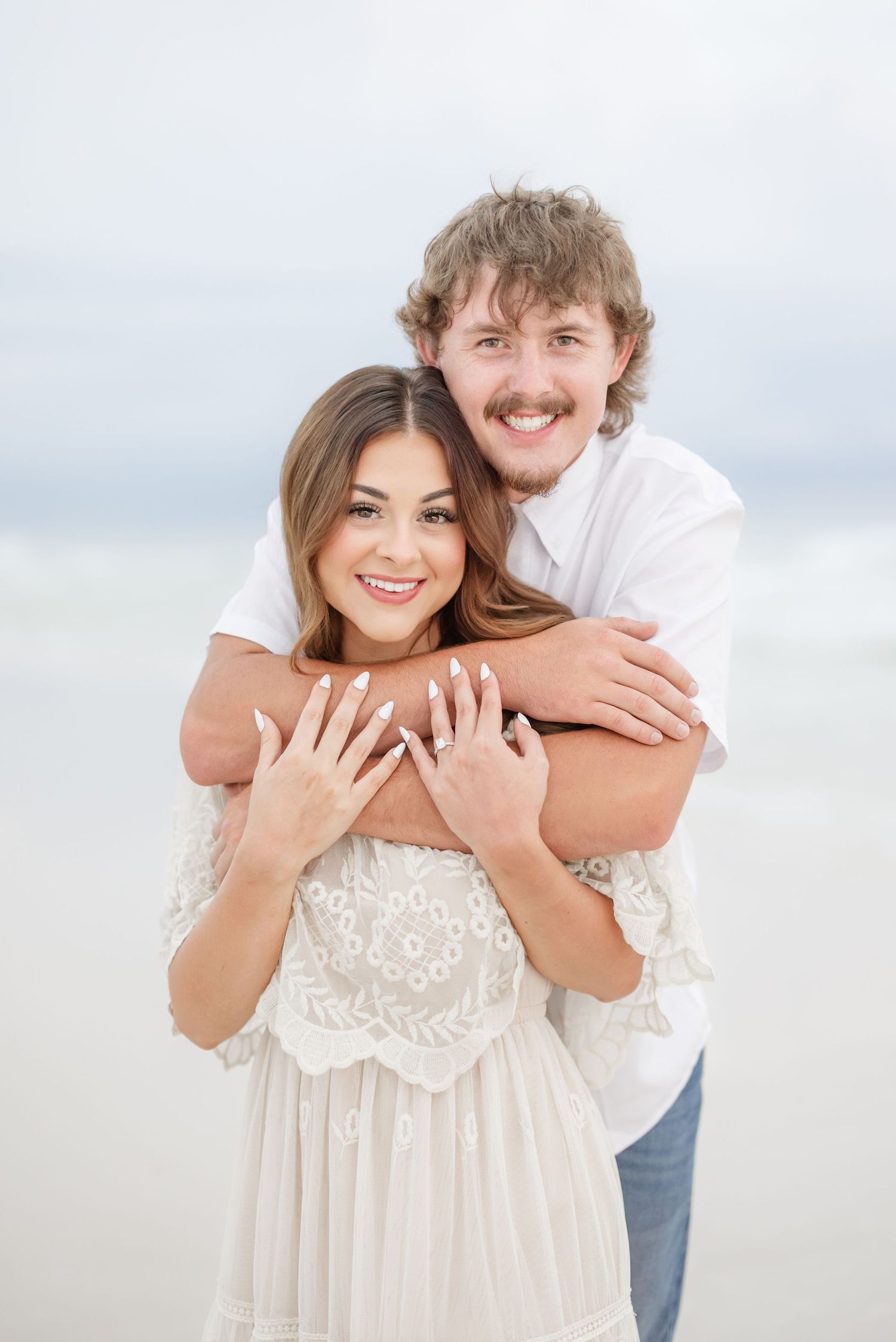 Couple embraces on a bright beach in matching white outfits during a romantic photoshoot.
