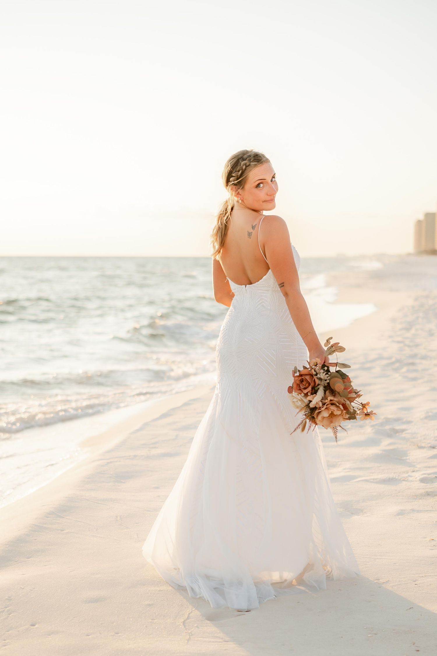A bride in a flowing white dress poses on a beach at sunset while holding a dried floral bouquet.