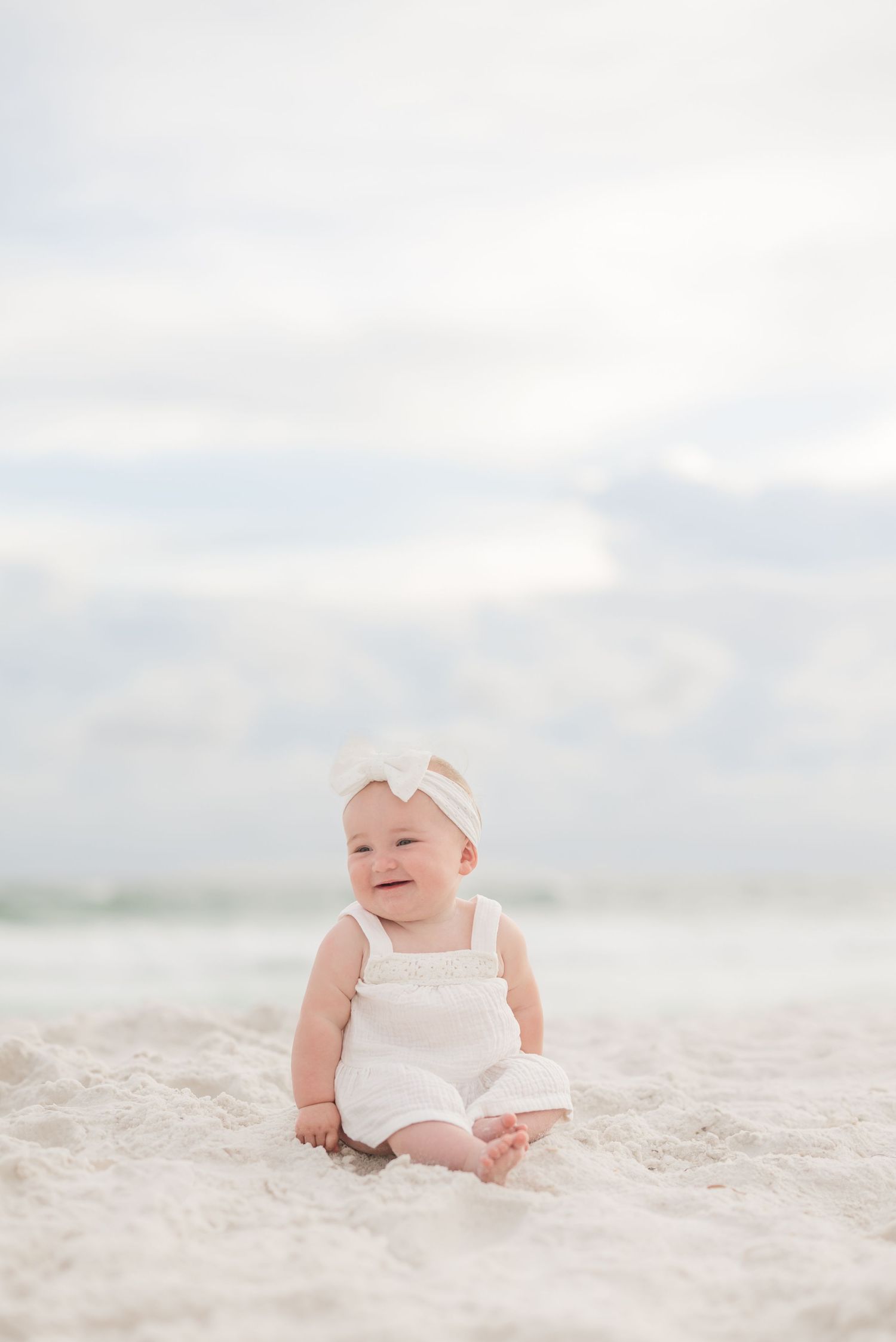 Baby in white romper sitting on sandy beach with cloudy sky background in Panama City Beach, Fl..