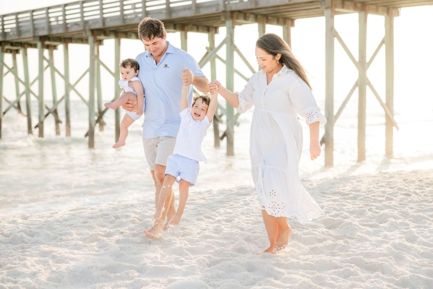 Family enjoying a sunset beach photoshoot near a wooden pier on white sand.