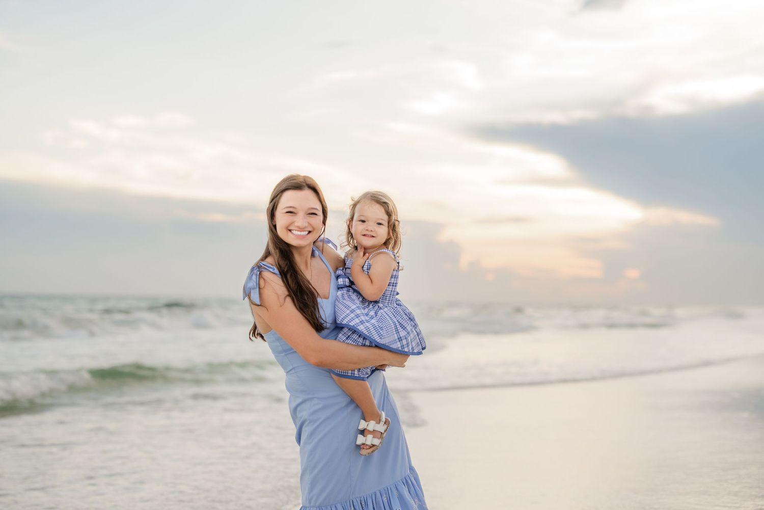 Mother and daughter in matching blue dresses share tender moments on a sunset beach.