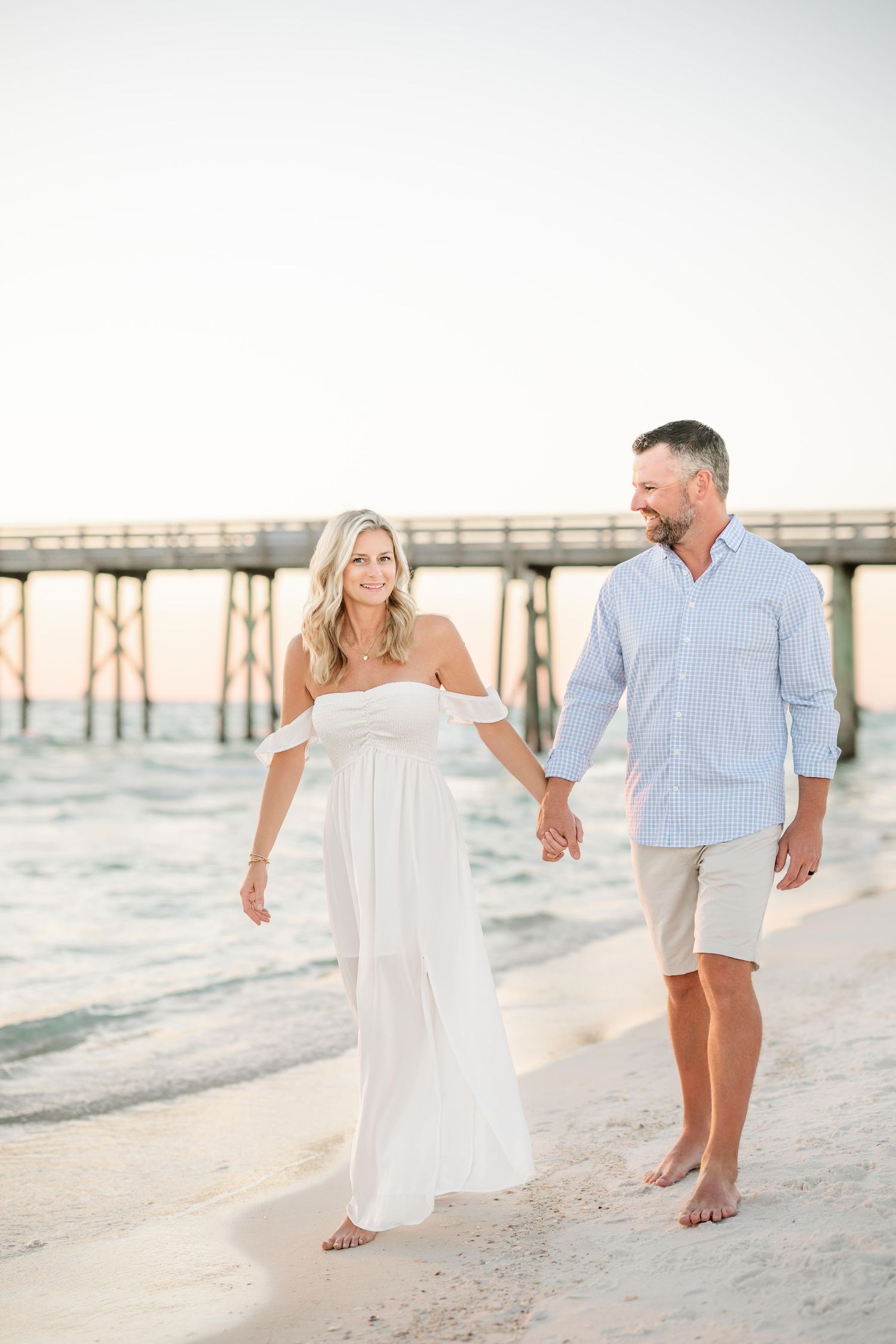 A couple strolls barefoot on the beach with a wooden pier in the background during sunset.