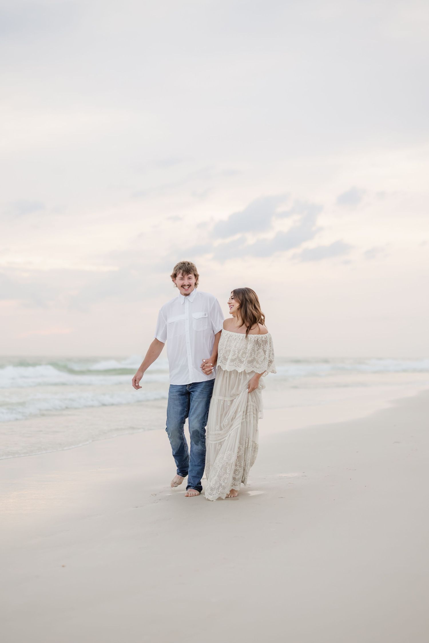 Couple walking along pristine beach shoreline in ethereal white dress and casual wear.