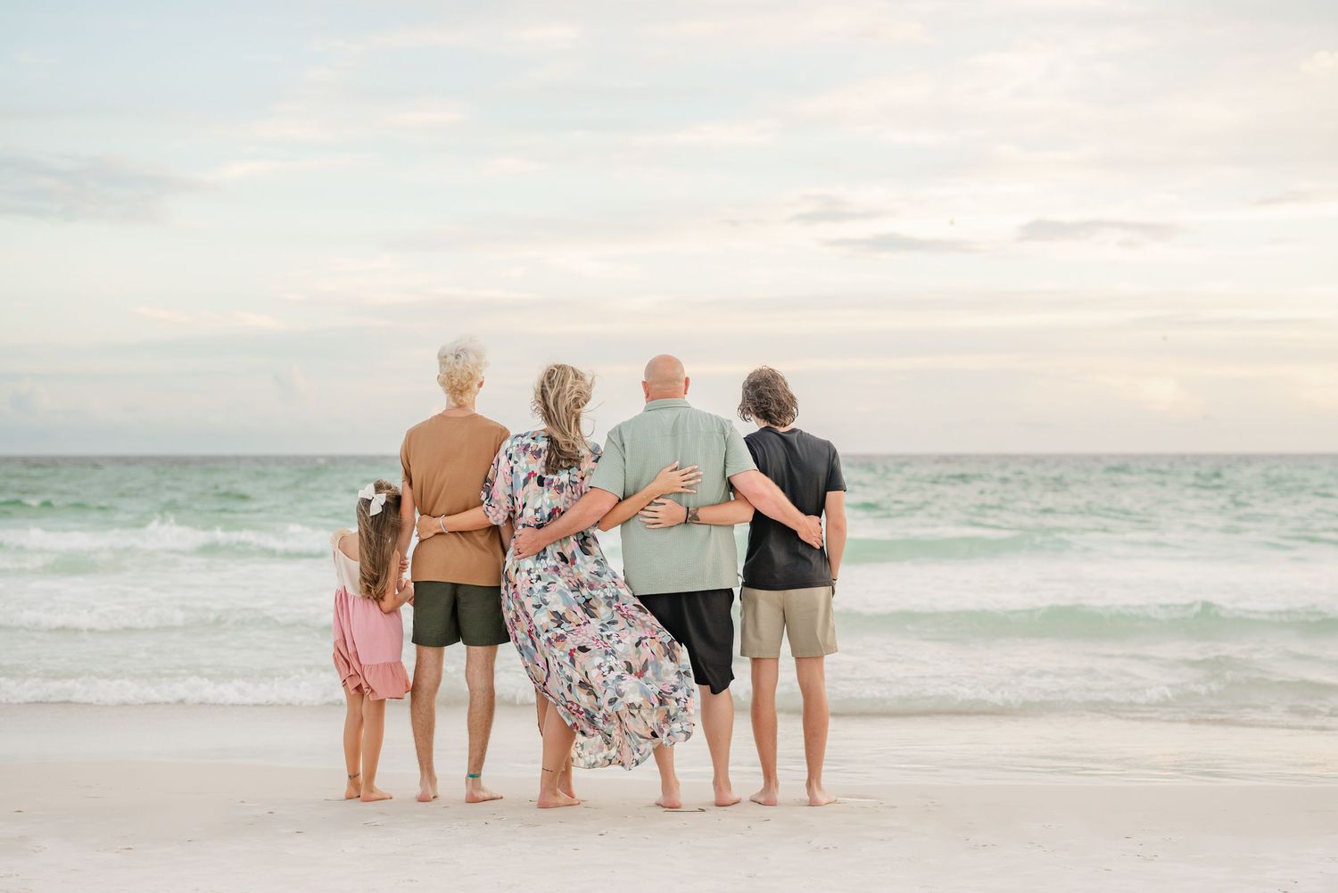 Extended family stands together facing the ocean at sunset on a white sandy beach.