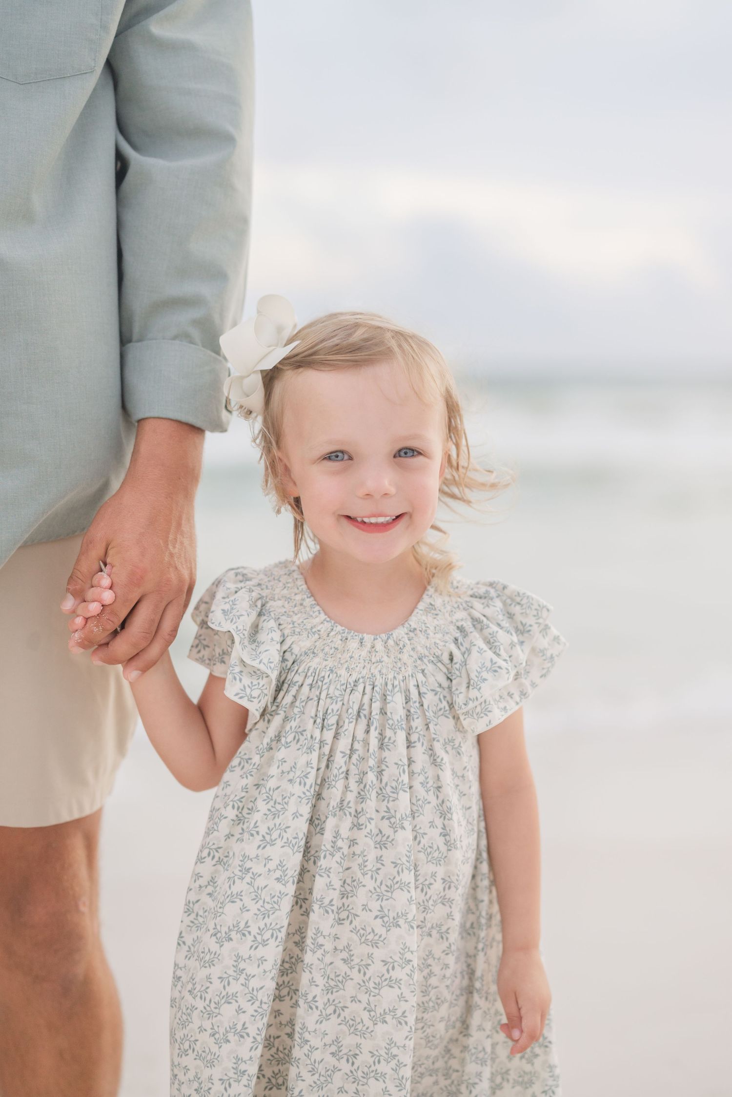 Child in floral dress smiling during beach photoshoot with family.
