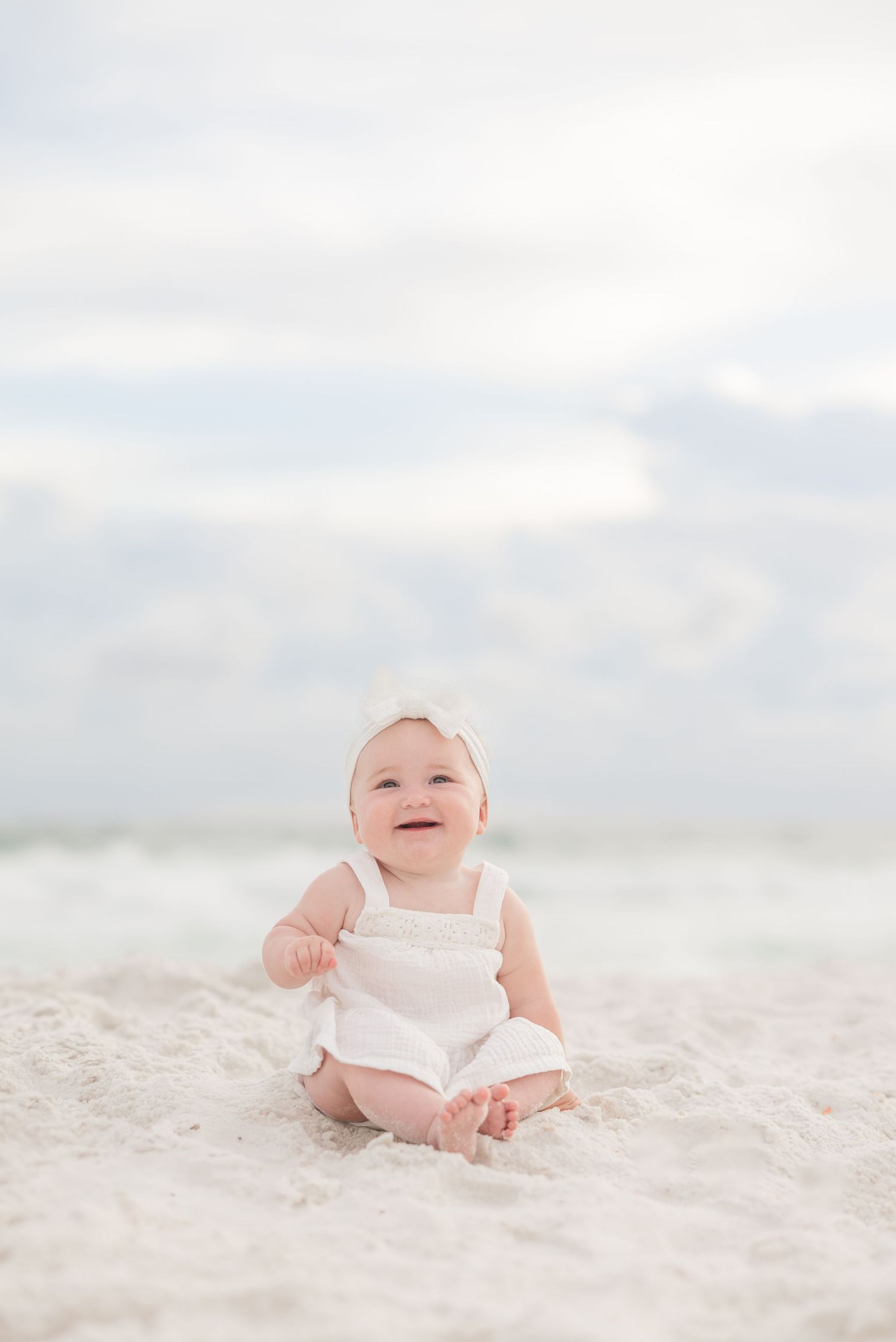 A baby in white clothing sits on a sunny beach with soft sand and cloudy sky in the background in Panama City Beach, Fl.
