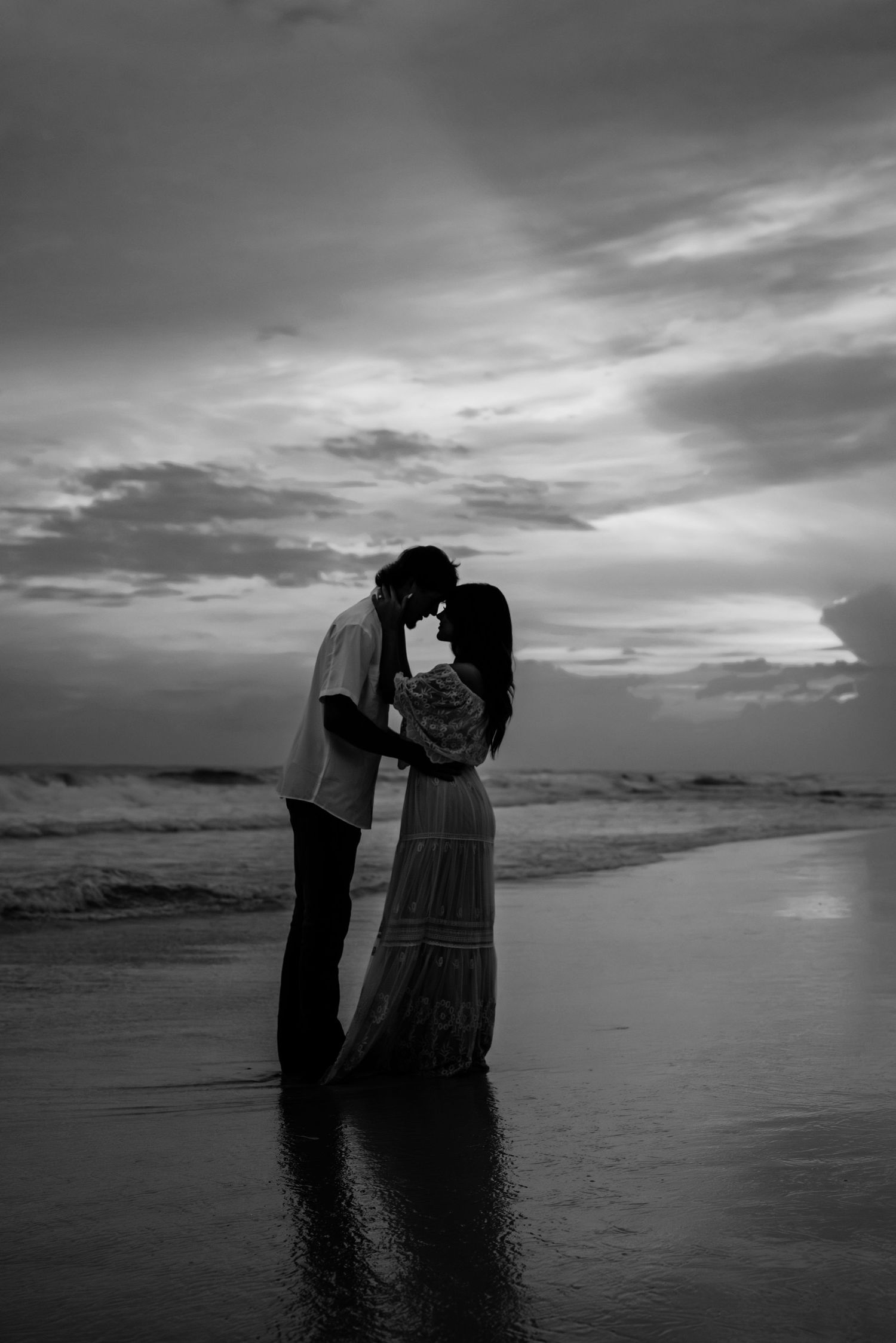 Black and white beach portrait capturing intimate silhouettes against dramatic sky.