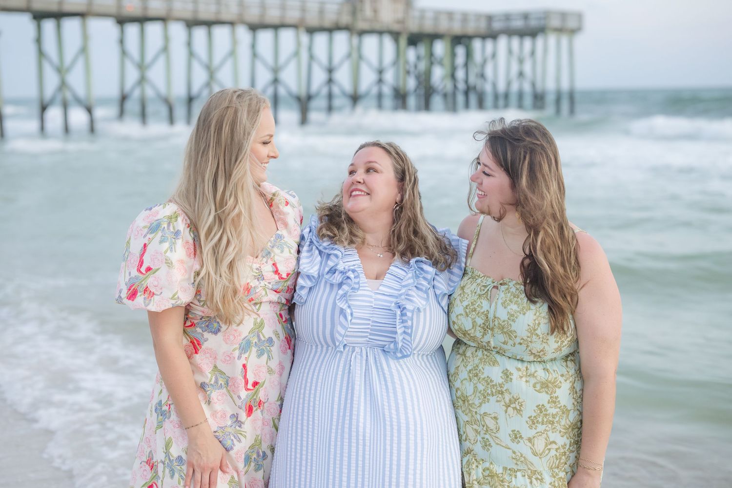 Women in floral and striped dresses sharing laughter on a breezy beach.