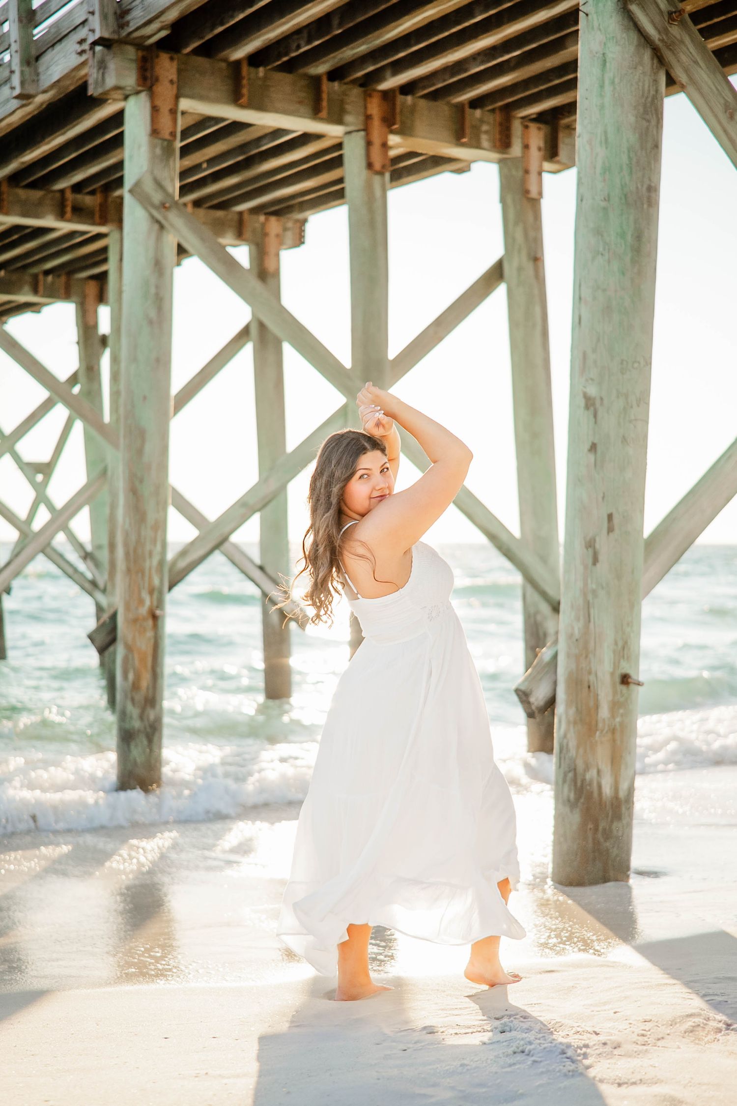 A dreamy beach photoshoot under a wooden pier with soft sunlight filtering through the slats.